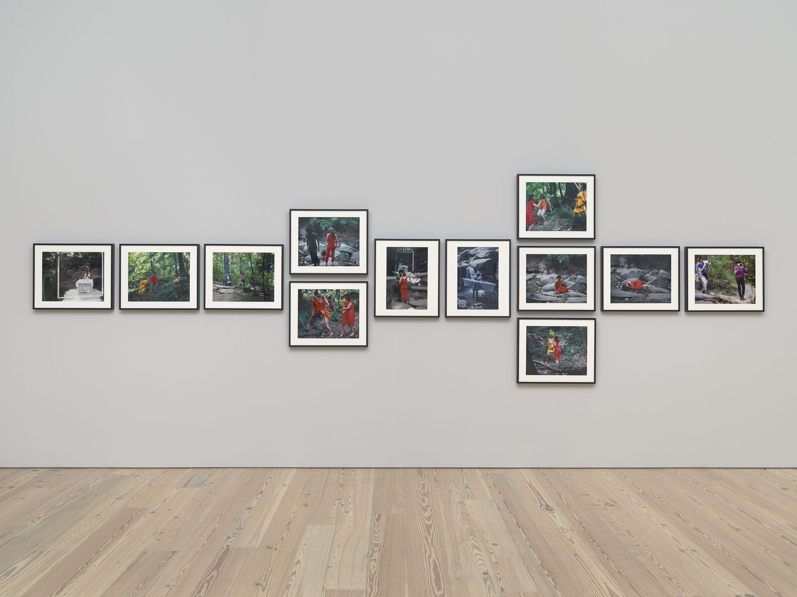 Twelve framed photographs on a gallery wall show people in colorful clothing walking and interacting in a forest.