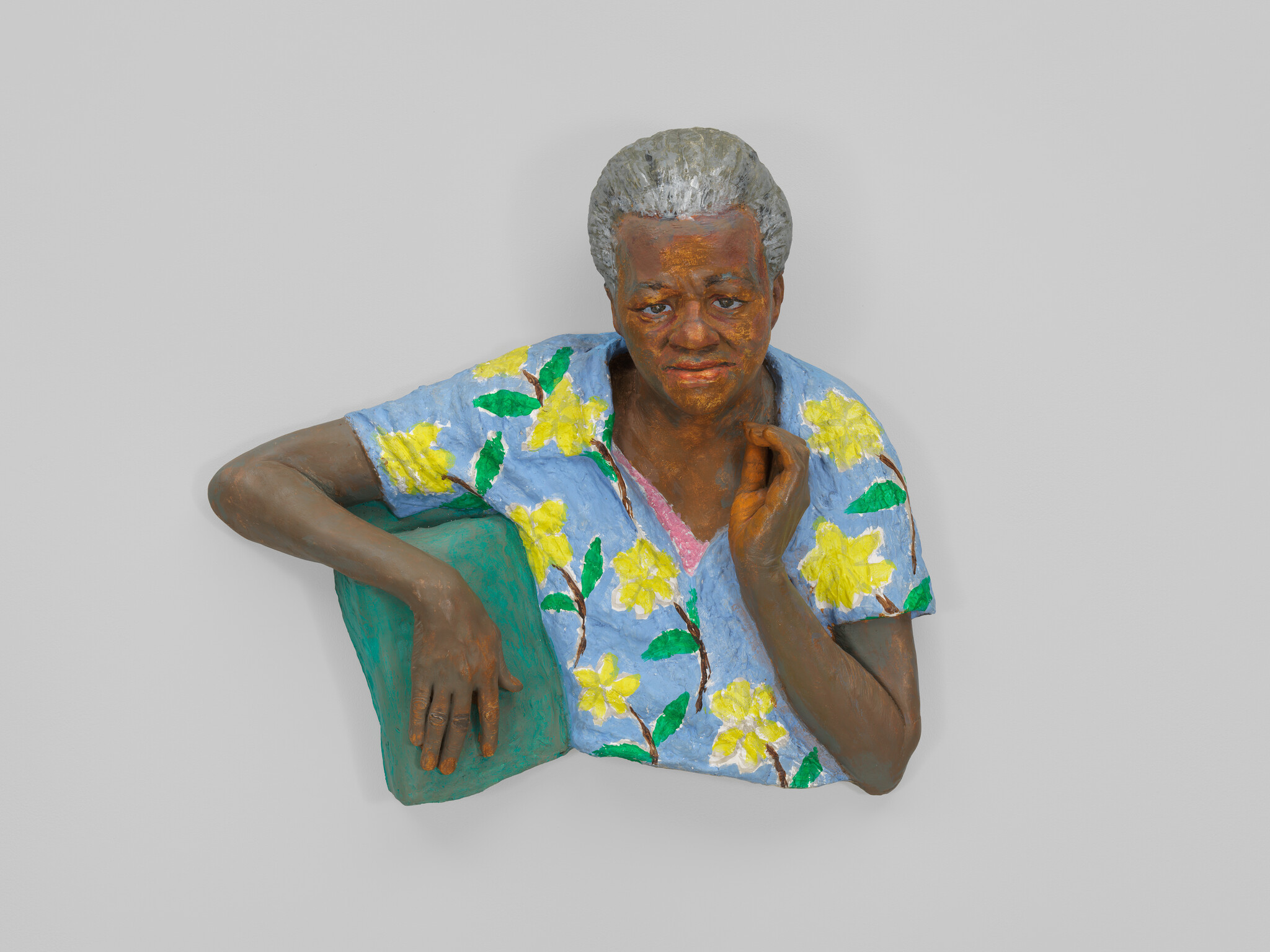 Elderly woman sculpture wearing a blue floral shirt, resting one arm on a green cushion.