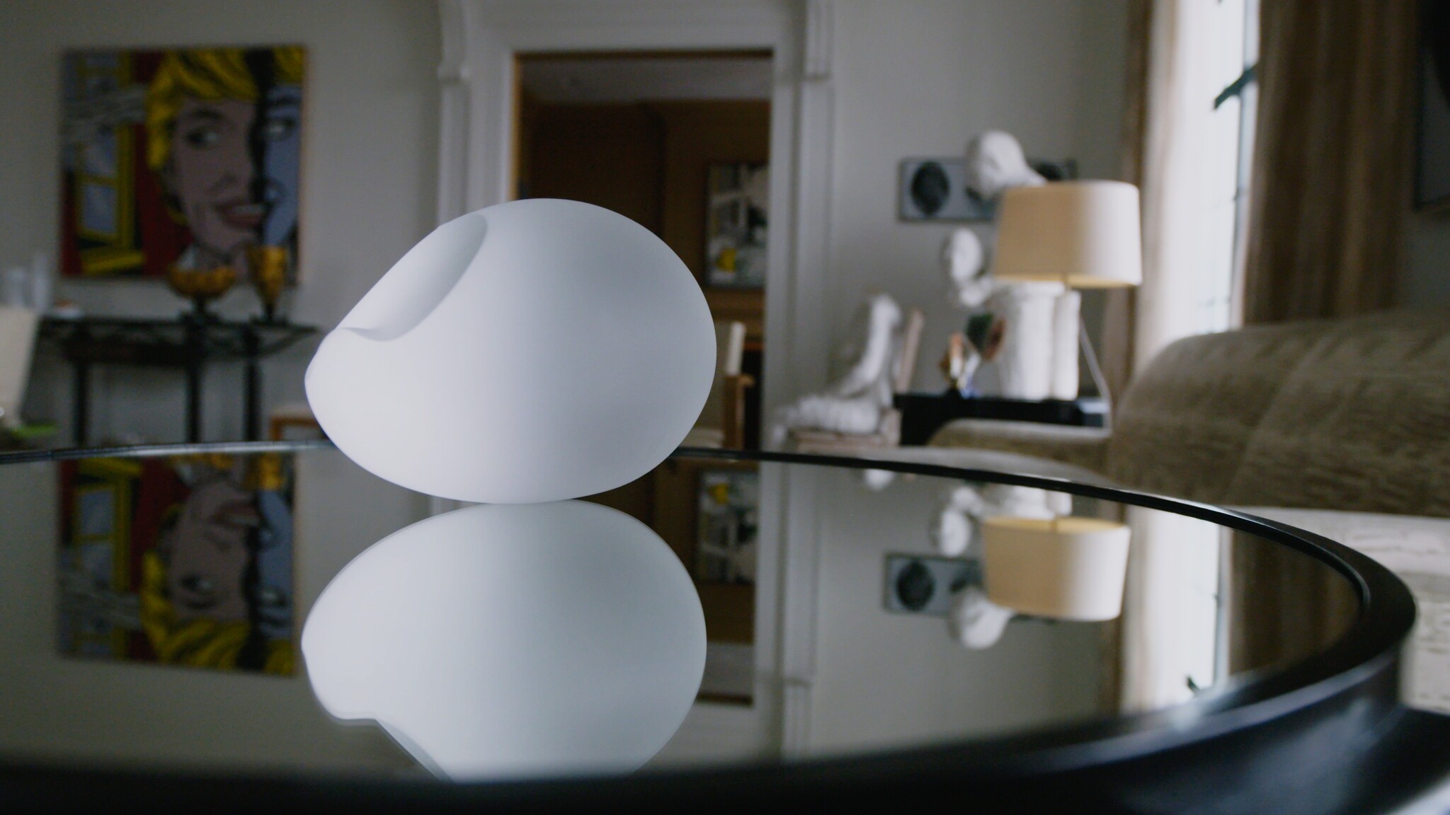 A smooth white decorative vase sits on a glass table, its reflection visible below.