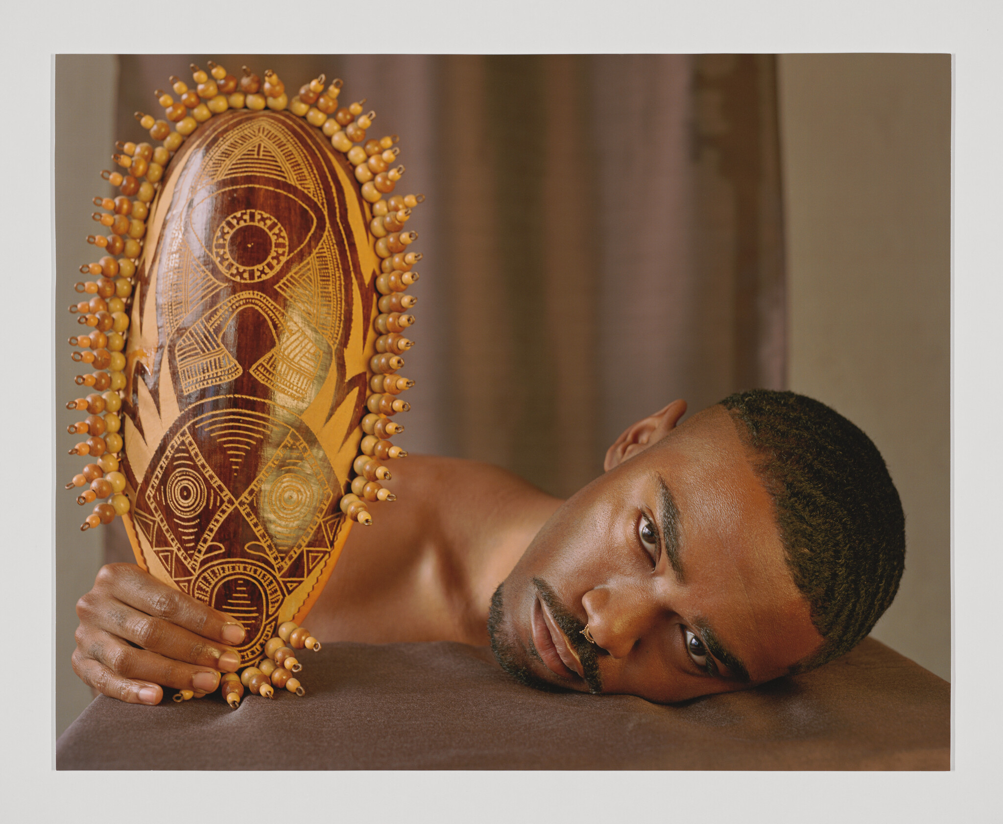 A Black man lies his head on a brown cloth in the foreground, holding an etched and beaded African mask