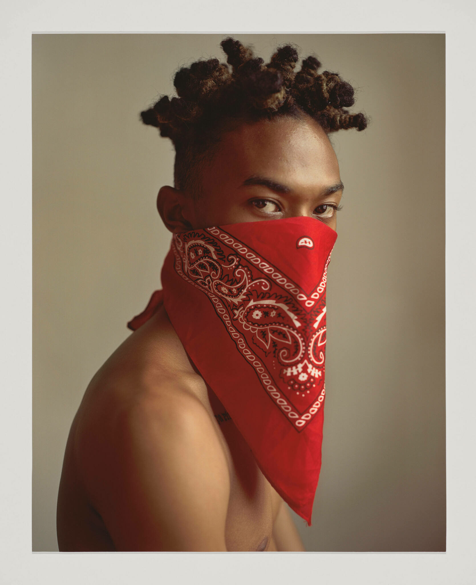 A young Black man stares at the viewer and red handkerchief tied around his head covers his nose and mouth 