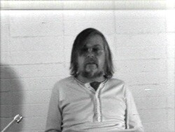 A man with shoulder-length hair and a beard sits against a plain white brick wall, looking forward.