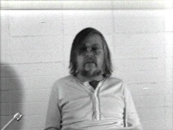 A bearded man with shoulder-length hair sits against a tiled wall.