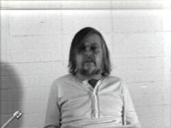 A bearded man with shoulder-length hair sits against a tiled wall.