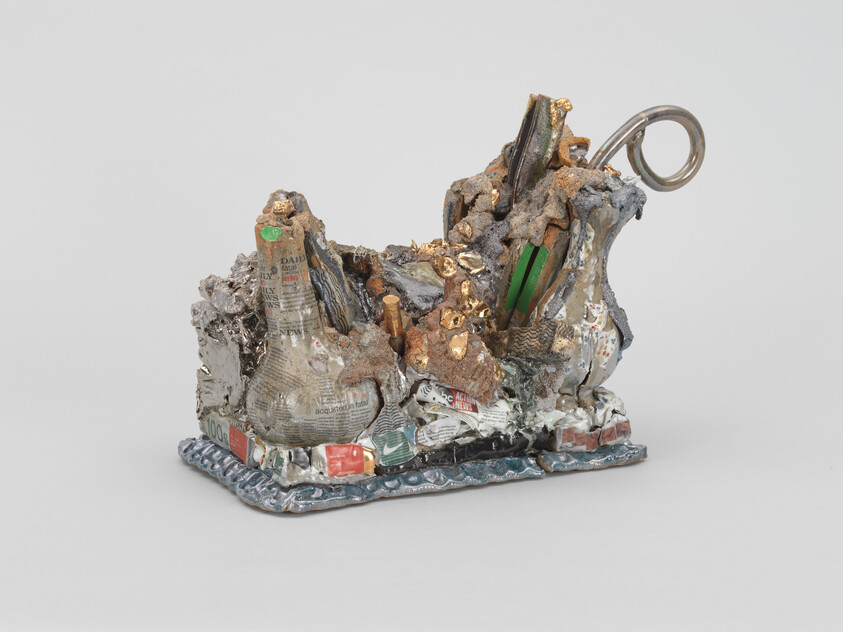 Small mixed-media sculpture made from crumpled newspaper, metal fragments, and gold-colored accents on a base.