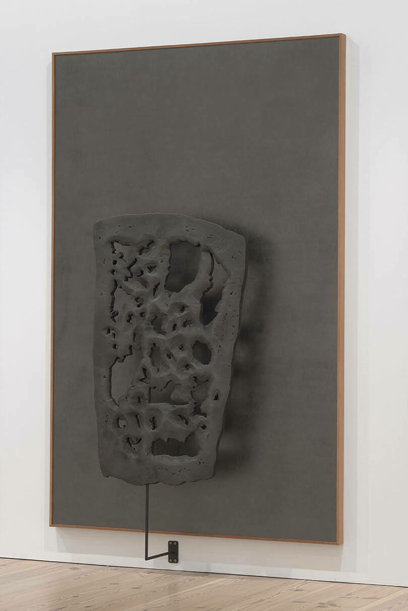 A perforated and craggy three-dimensional sculpture seems to hover in front of a sharply rectangular dark gray canvas.
