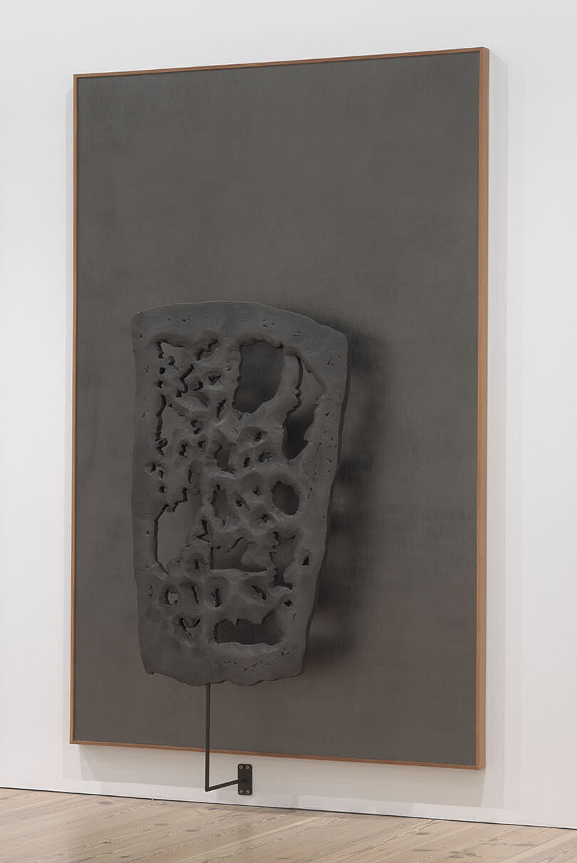 A perforated and craggy three-dimensional sculpture seems to hover in front of a sharply rectangular dark gray canvas.