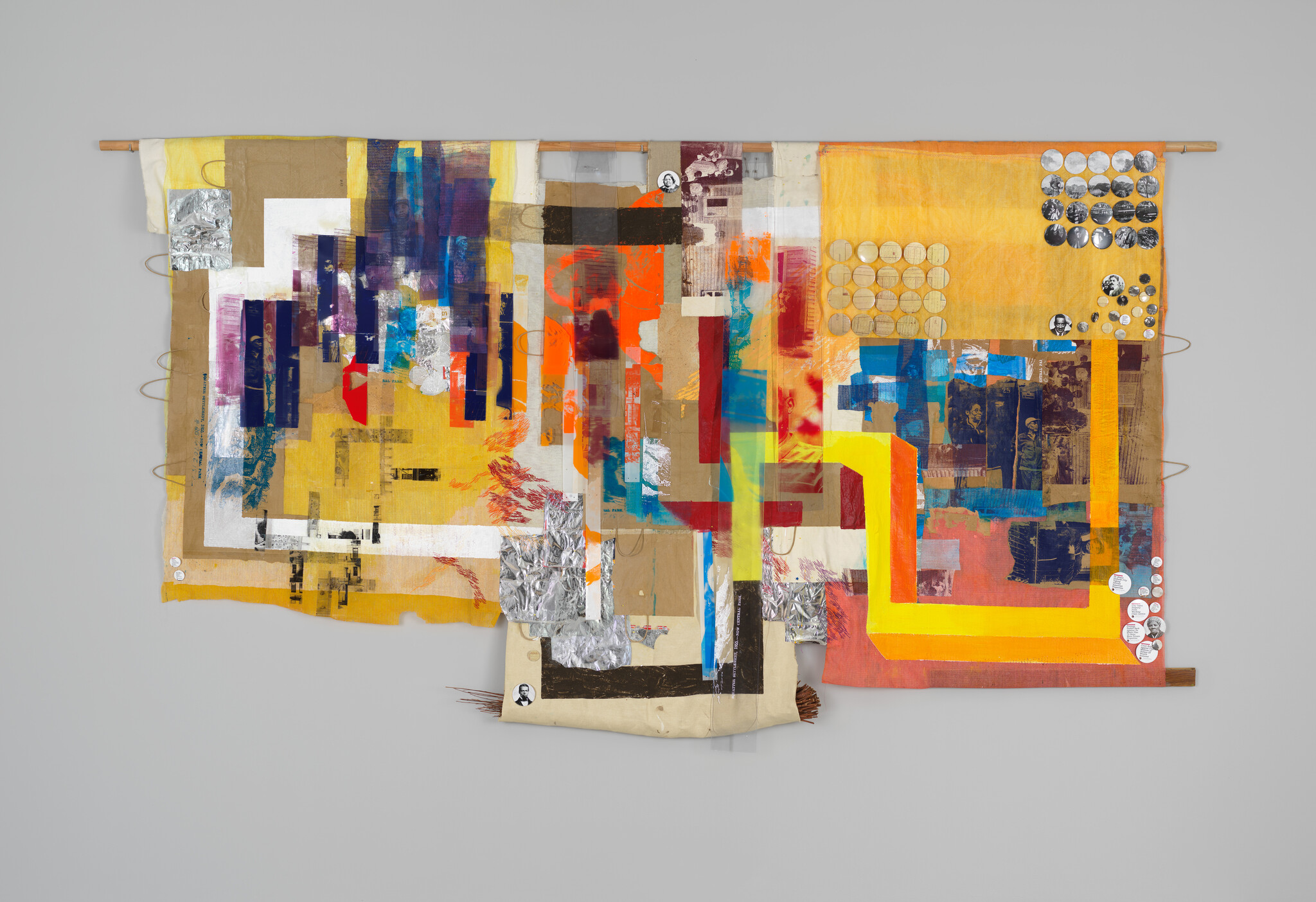 A vibrant mixed-media tapestry hangs against a light gray background, featuring an array of colors and materials. The artwork is composed of various fabrics and papers, with patches of yellow, orange, red, blue, and silver. Some sections have printed patterns or text, while others are painted or dyed. The piece is adorned with circular elements that resemble coins or buttons, creating a rich texture and depth. The tapestry is suspended from a horizontal wooden dowel, suggesting a blend of traditional craft and contemporary art.
