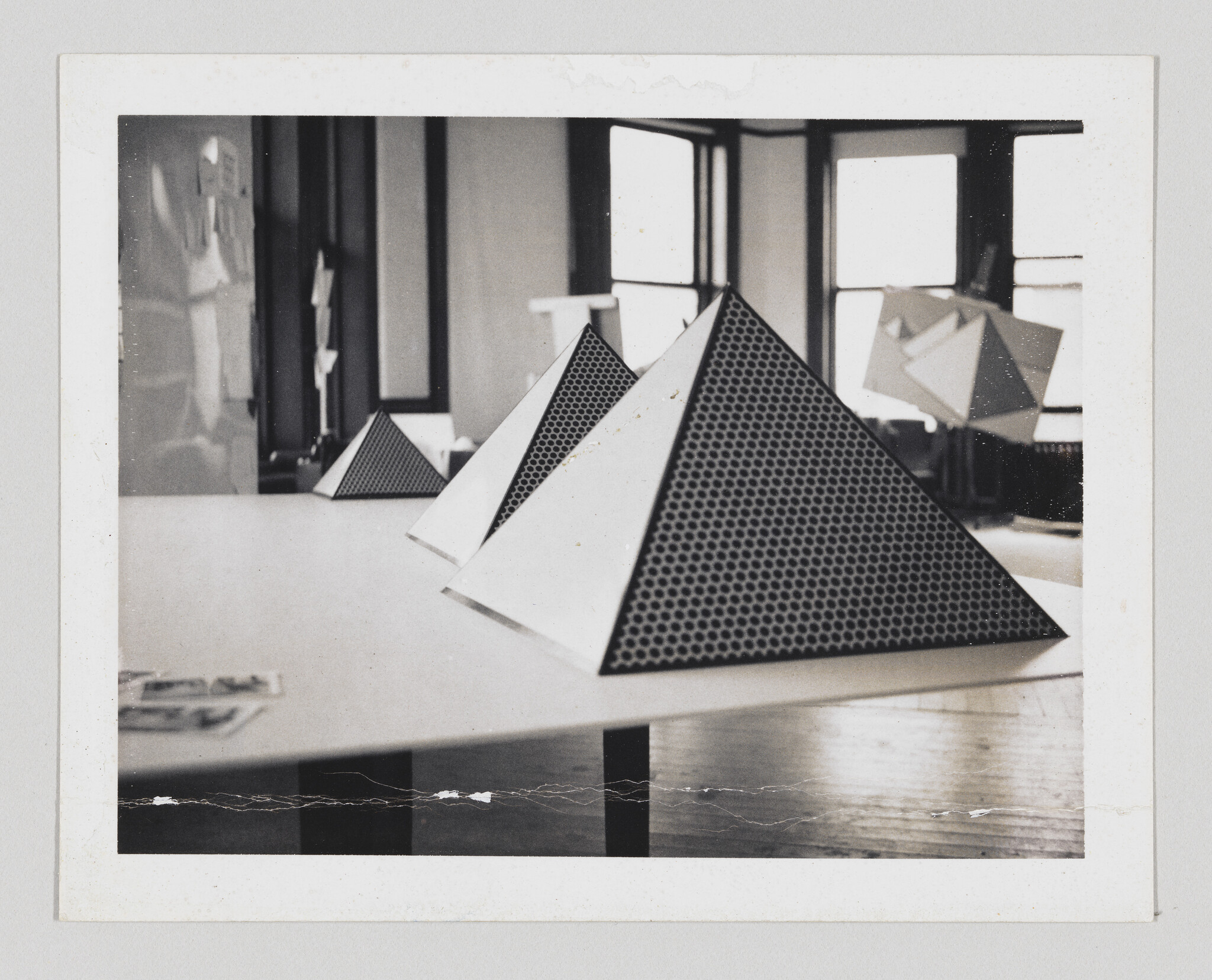 A vintage-looking photograph depicting several geometric models, possibly made of paper or light metal, with a perforated pattern. The models are placed on a table in a room with large windows, and there's a reflection on the polished surface of the table. The photo has a white border, and it appears aged with some creases and discoloration.