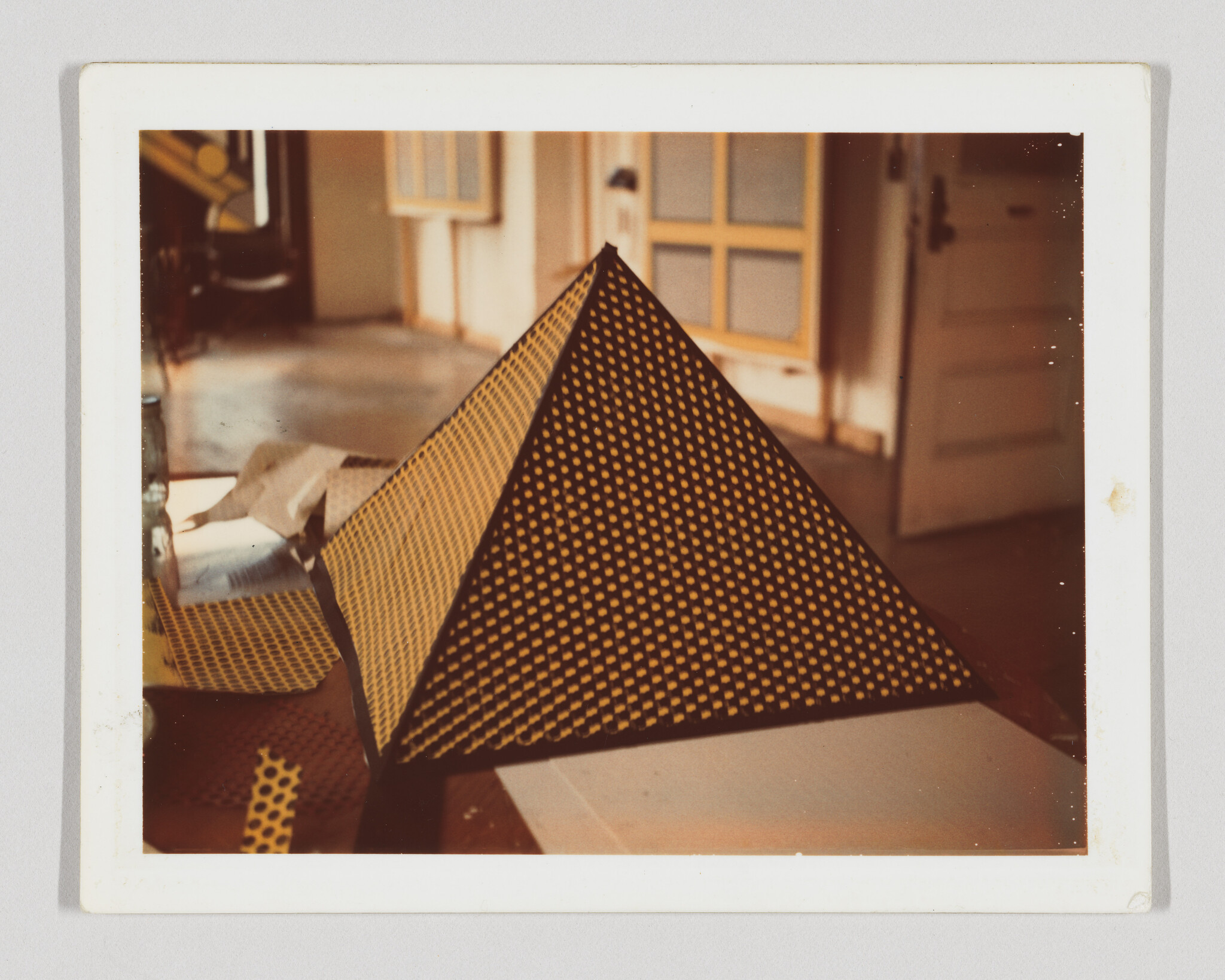 A black pyramid covered in yellow dot-patterned mesh sits on a cluttered table in a room.