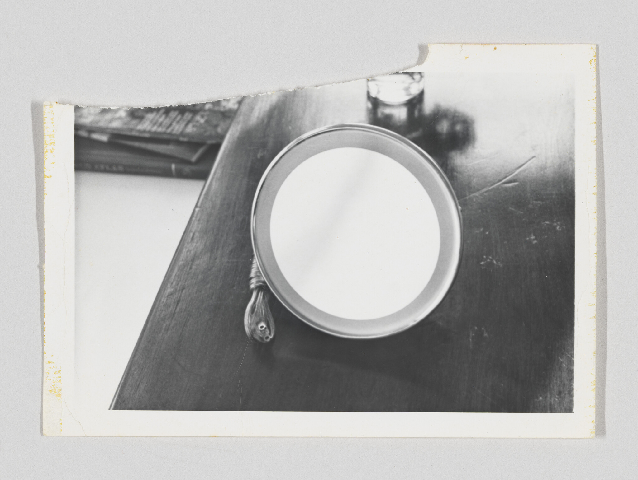 Round hand mirror with tassel lies face-up on a dark wooden table near a drinking glass.