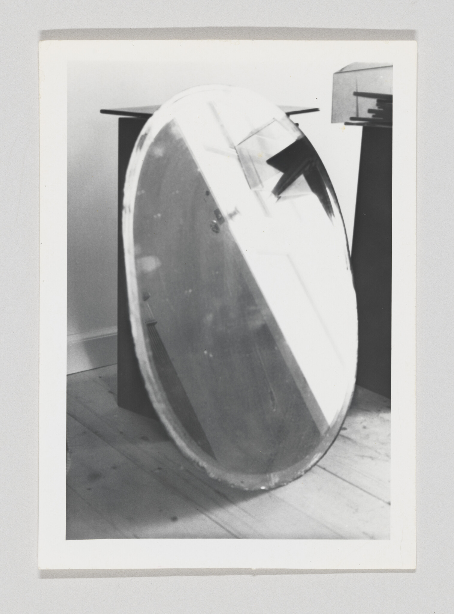 A black and white photograph showing a large, round, metallic object leaning against a wall in an indoor setting. The object appears to be a reflective disc with some tape or attachments on its surface. It is positioned on a wooden floor near a table or desk that is partially visible in the background.