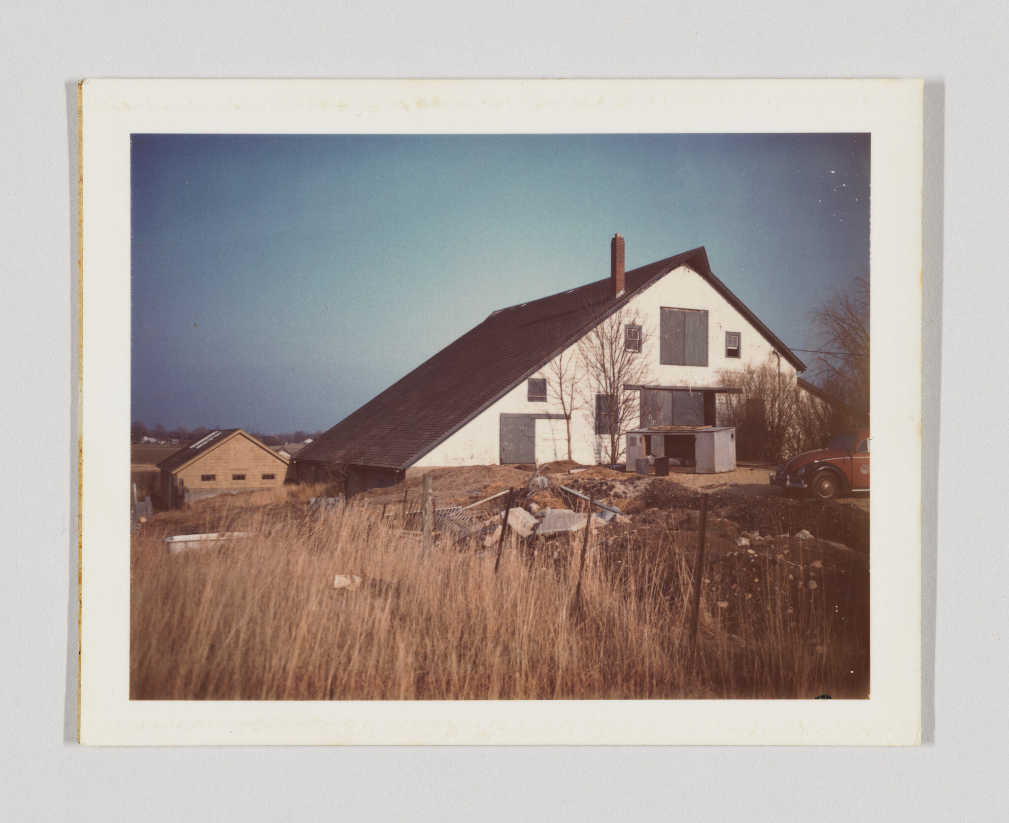 A large white barn with a steep dark roof sits behind tall dry grass and a parked car.