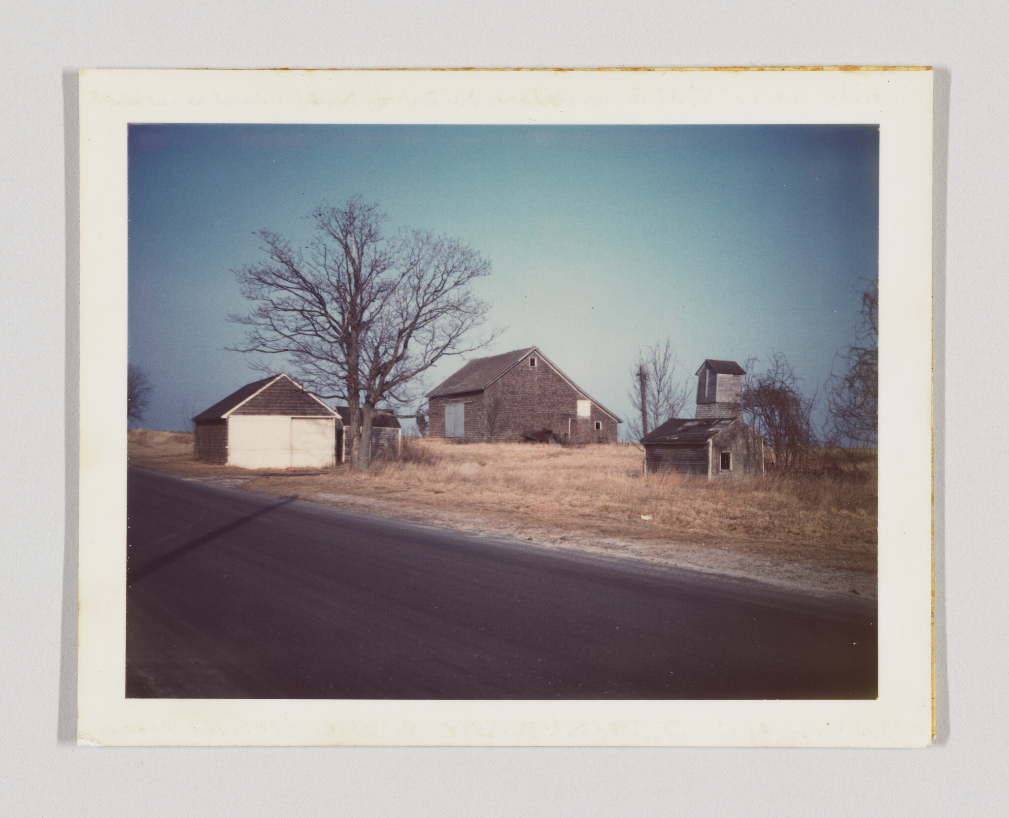 Abandoned barns and a small shed stand in dry grass beside a quiet country road.