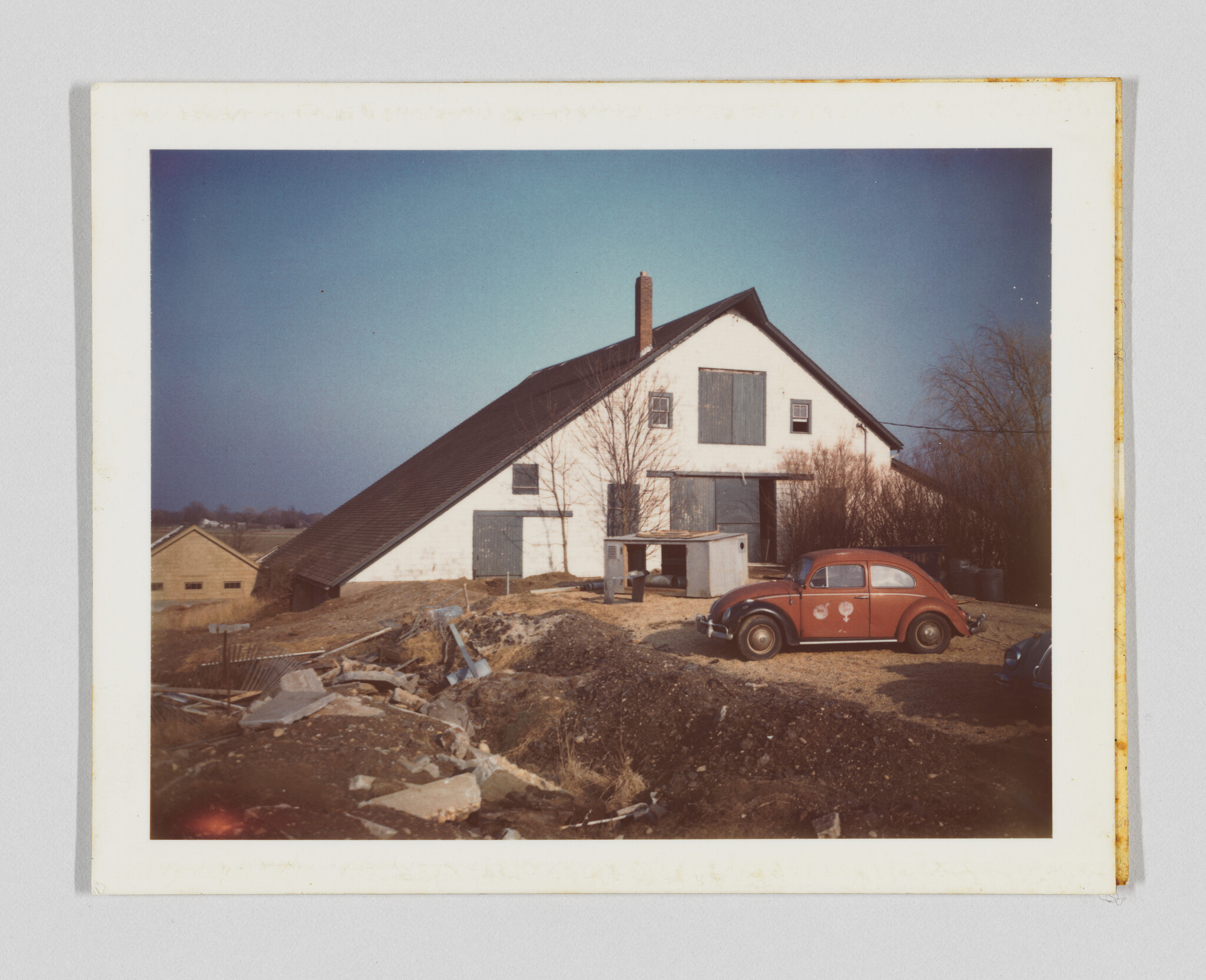 A white barn with a steep roof stands behind a parked red Volkswagen Beetle in a yard.
