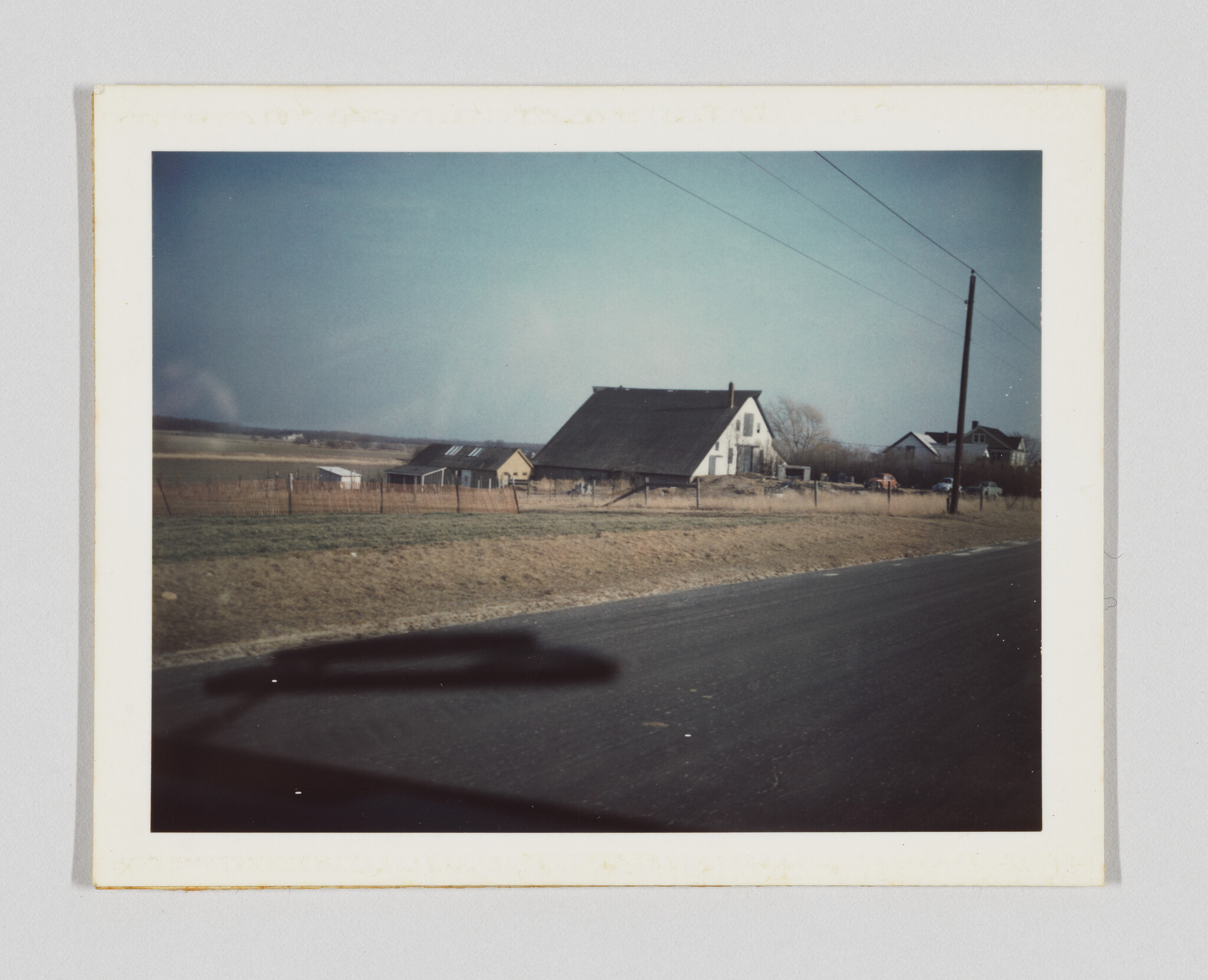 A large dark-roofed barn and farmhouse sit behind a fence beside a quiet country road.