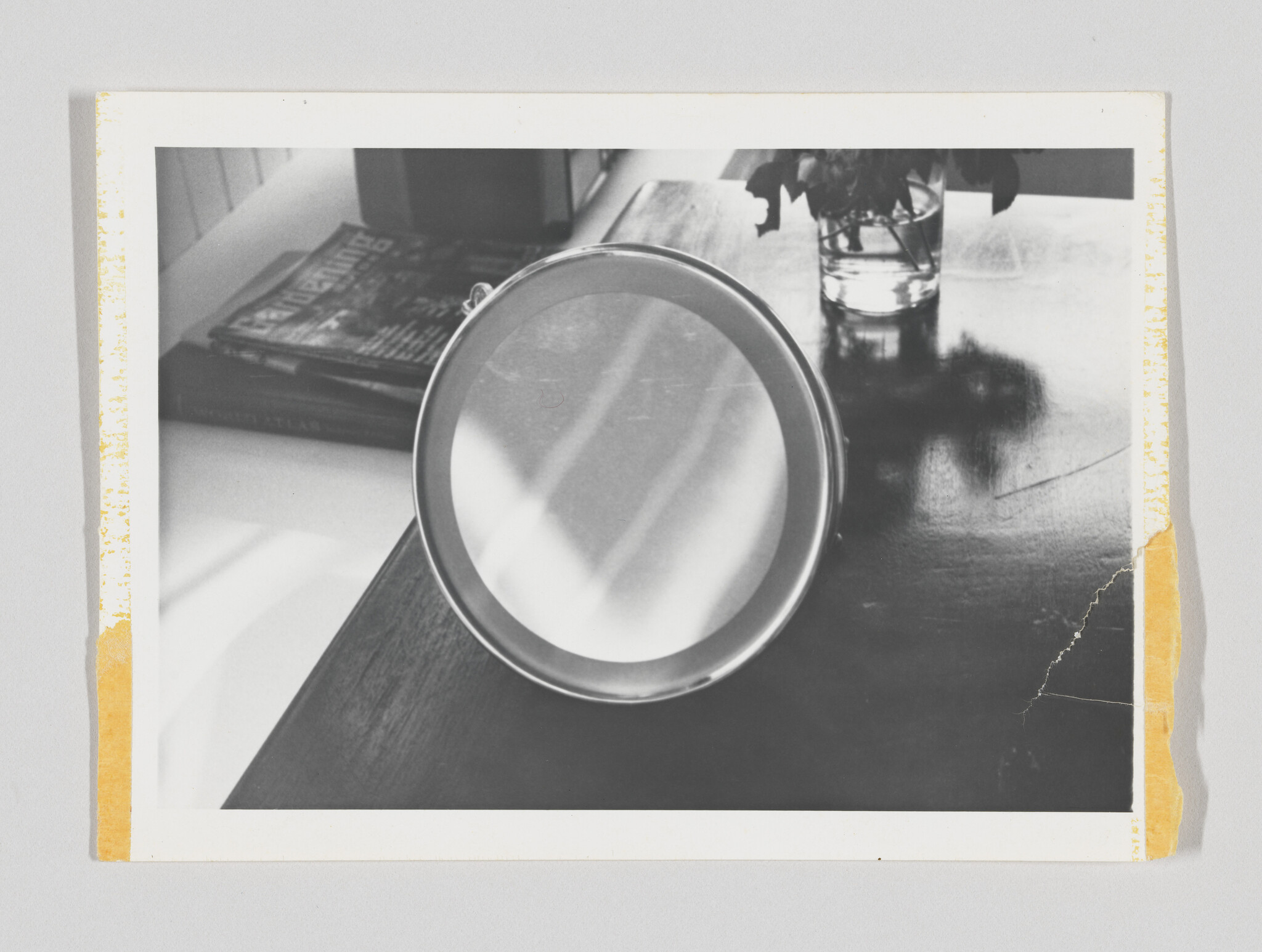 A round handheld mirror rests on a wooden table near magazines and a small vase.