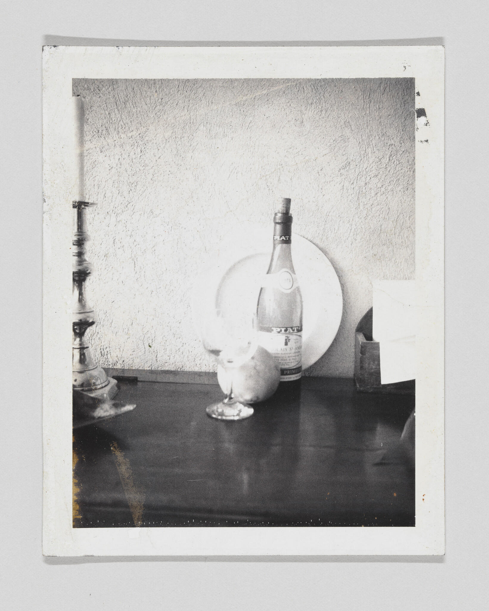 A black and white photograph depicting a still life scene with a bottle, a glass, and a candlestick on a dark surface against a textured wall. The photo has a white border and shows signs of age with some discoloration and damage to the edges.