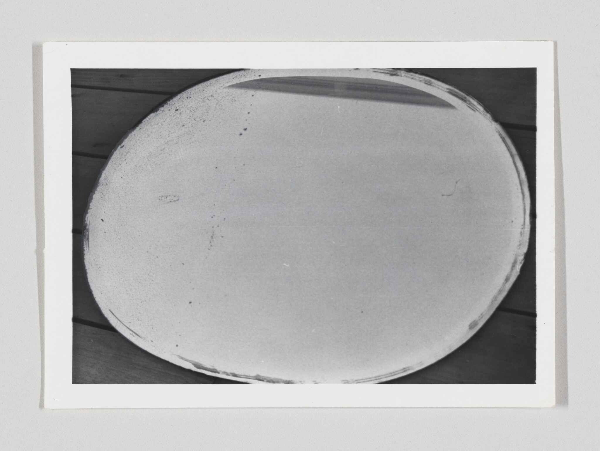 Large round blank plate resting on wooden table, photographed in black and white.
