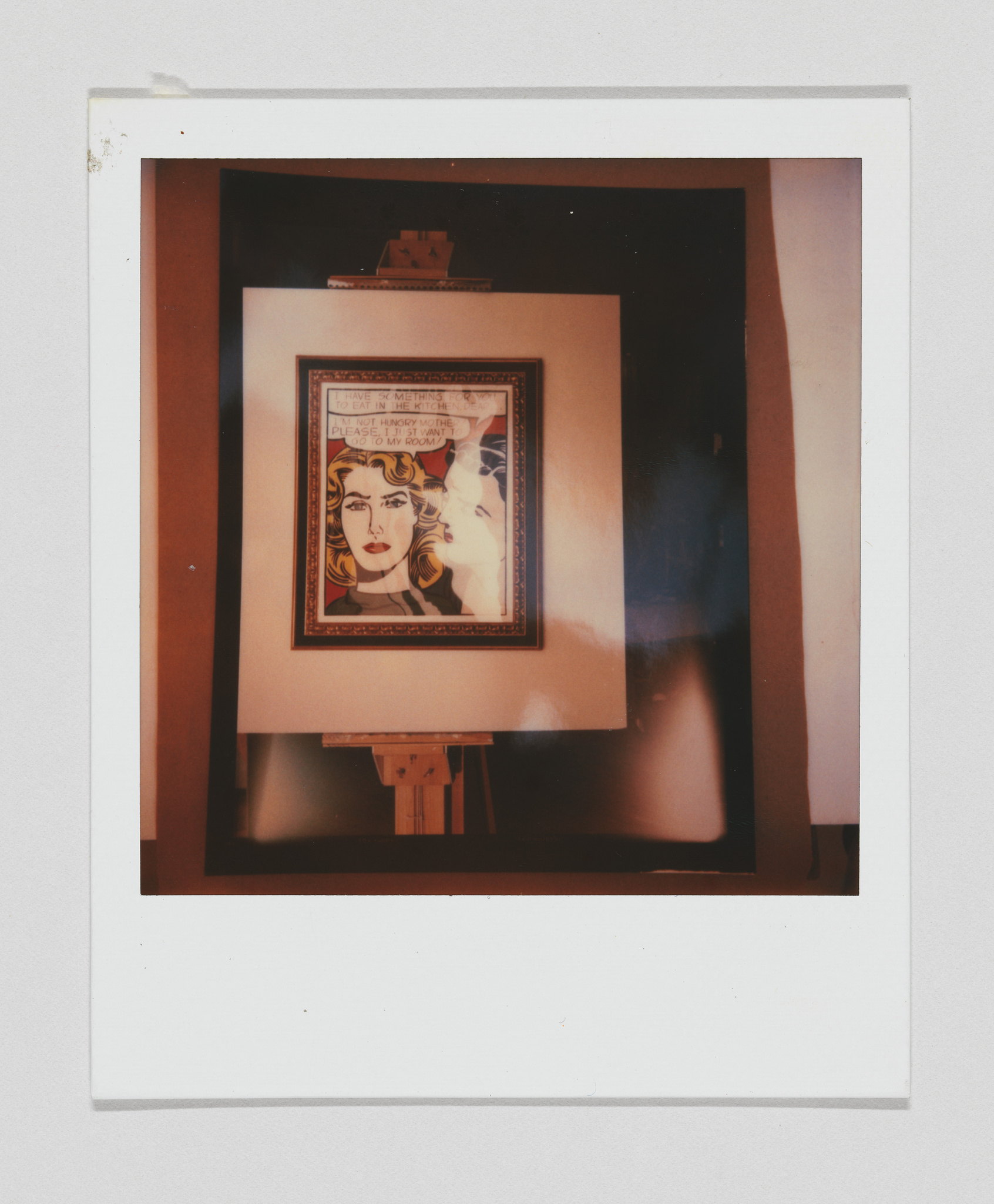 A Polaroid photo capturing a framed artwork on an easel, featuring a stylized portrait of a woman with text elements, set against a dark background.