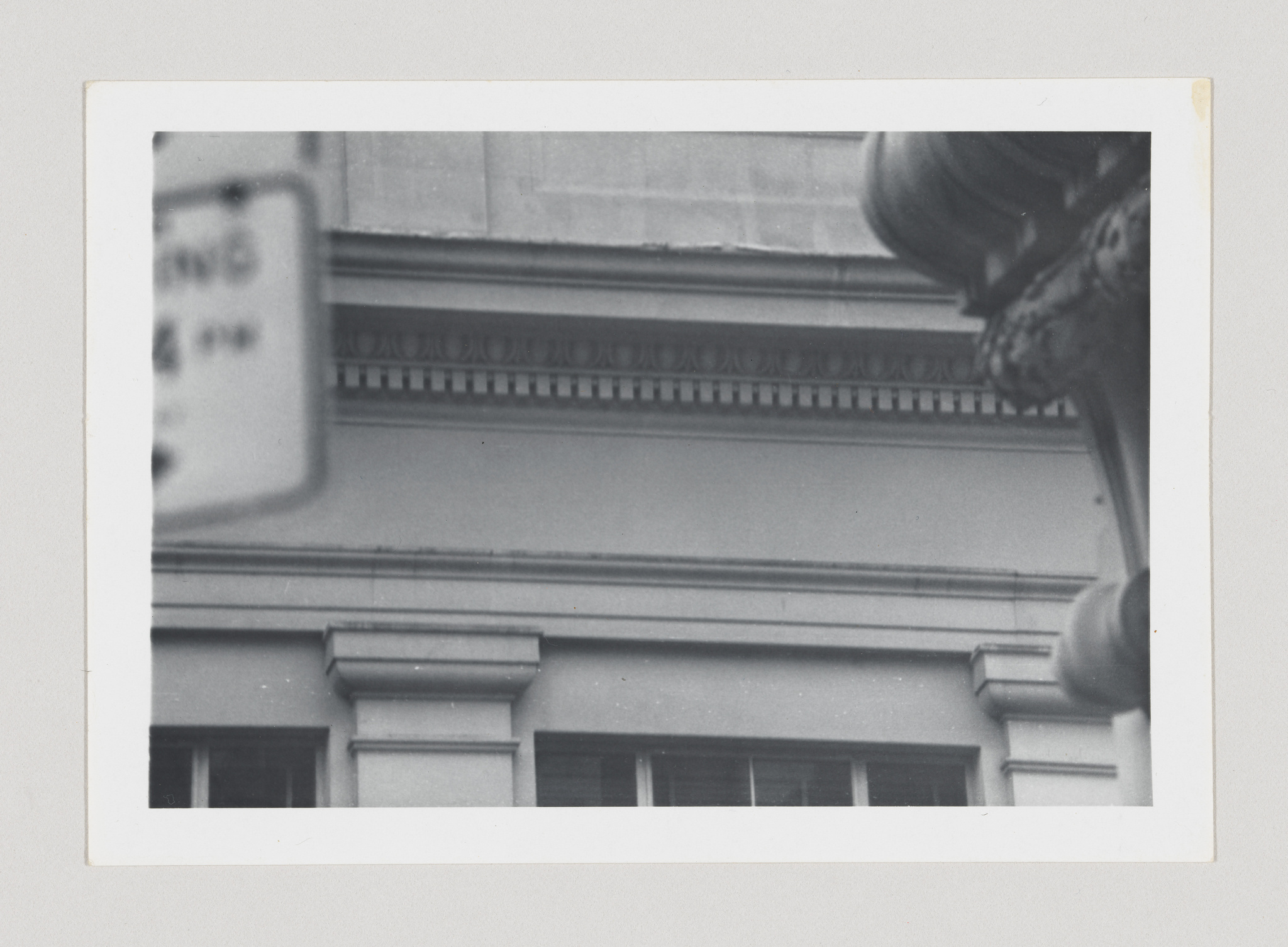 A black and white photograph showing a partial view of a building's classical architectural details, with a blurred street sign in the foreground to the left and a portion of a street lamp to the right. The image has a vintage appearance, with a white border framing the photo.