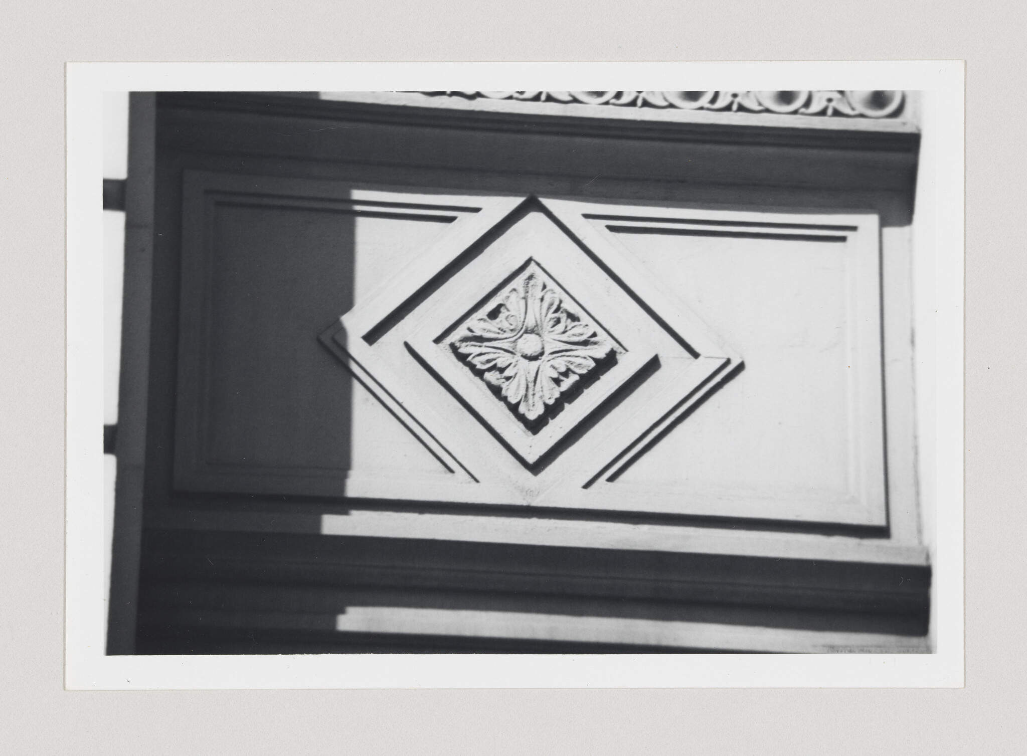Black and white photo of an architectural detail showing a diamond-shaped ornament with a leaf motif, set within a series of rectangular frames on a wall, casting shadows in the sunlight.