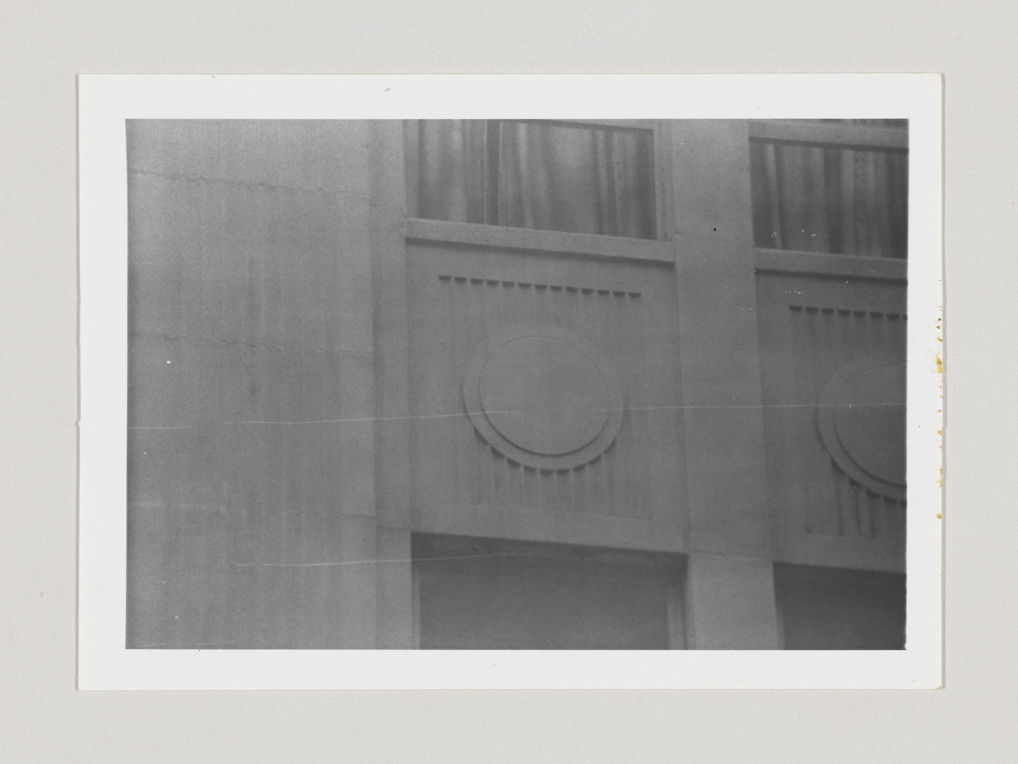 A black and white photograph showing a close-up of architectural details on a building, featuring two sections with decorative molding and circular motifs. The image has crease marks and signs of age, with the right edge slightly damaged, indicating it is an old photograph.