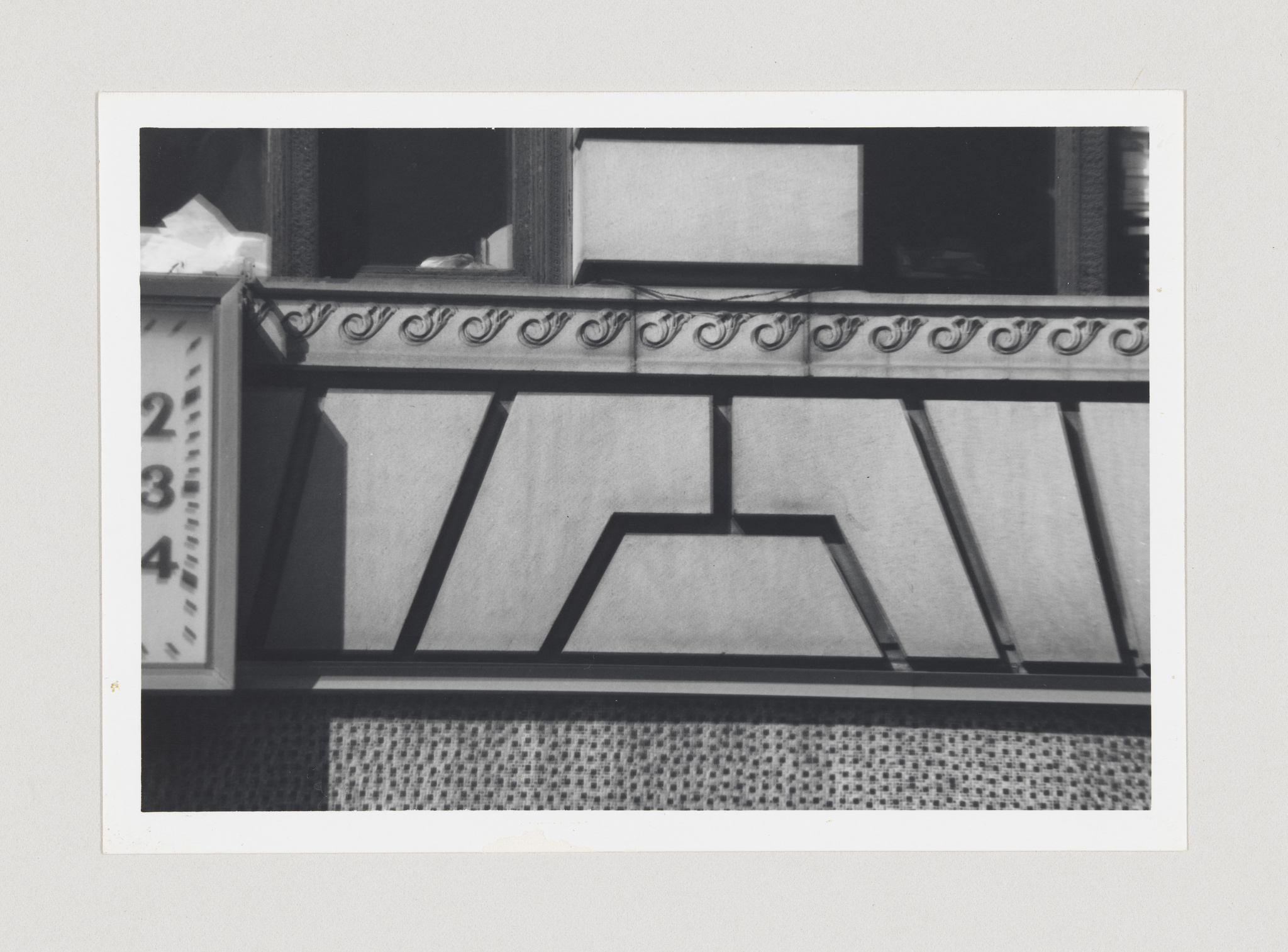 Black and white photo of a building's ornate balcony with decorative ironwork, featuring geometric patterns on the facade below the railing, and a partially visible window with papers on the sill.