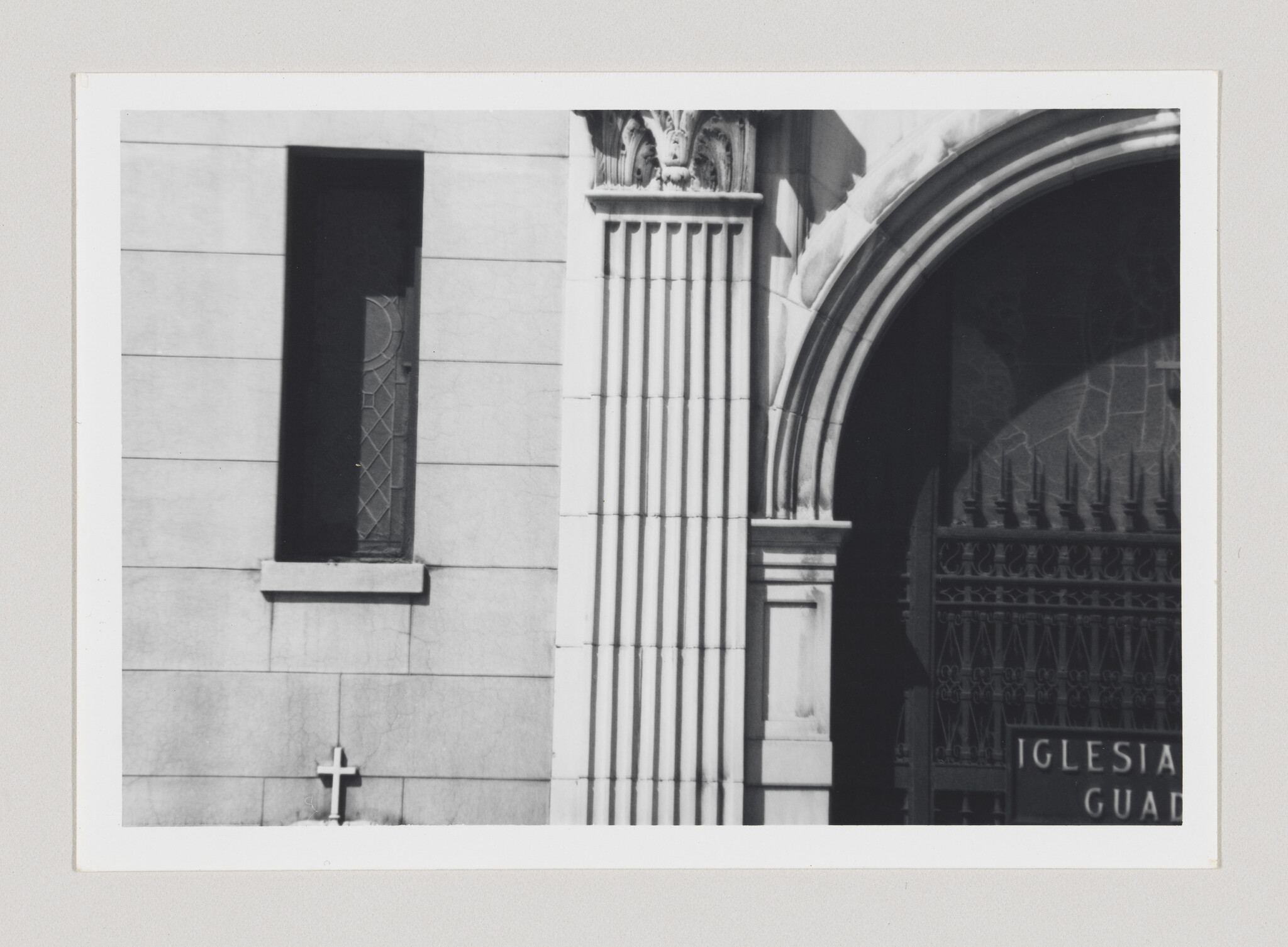 Stone church facade with narrow stained-glass window, fluted column, arched entrance, and small cross.