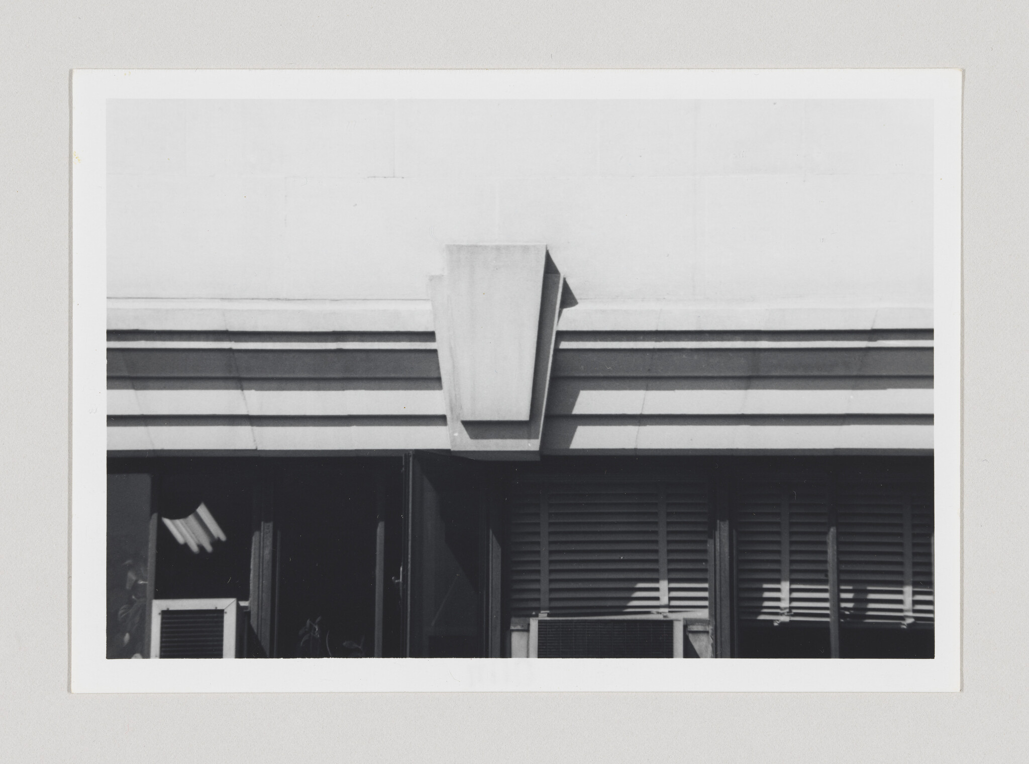 A building facade with layered cornice and a central projecting ornament above windows and air conditioners.