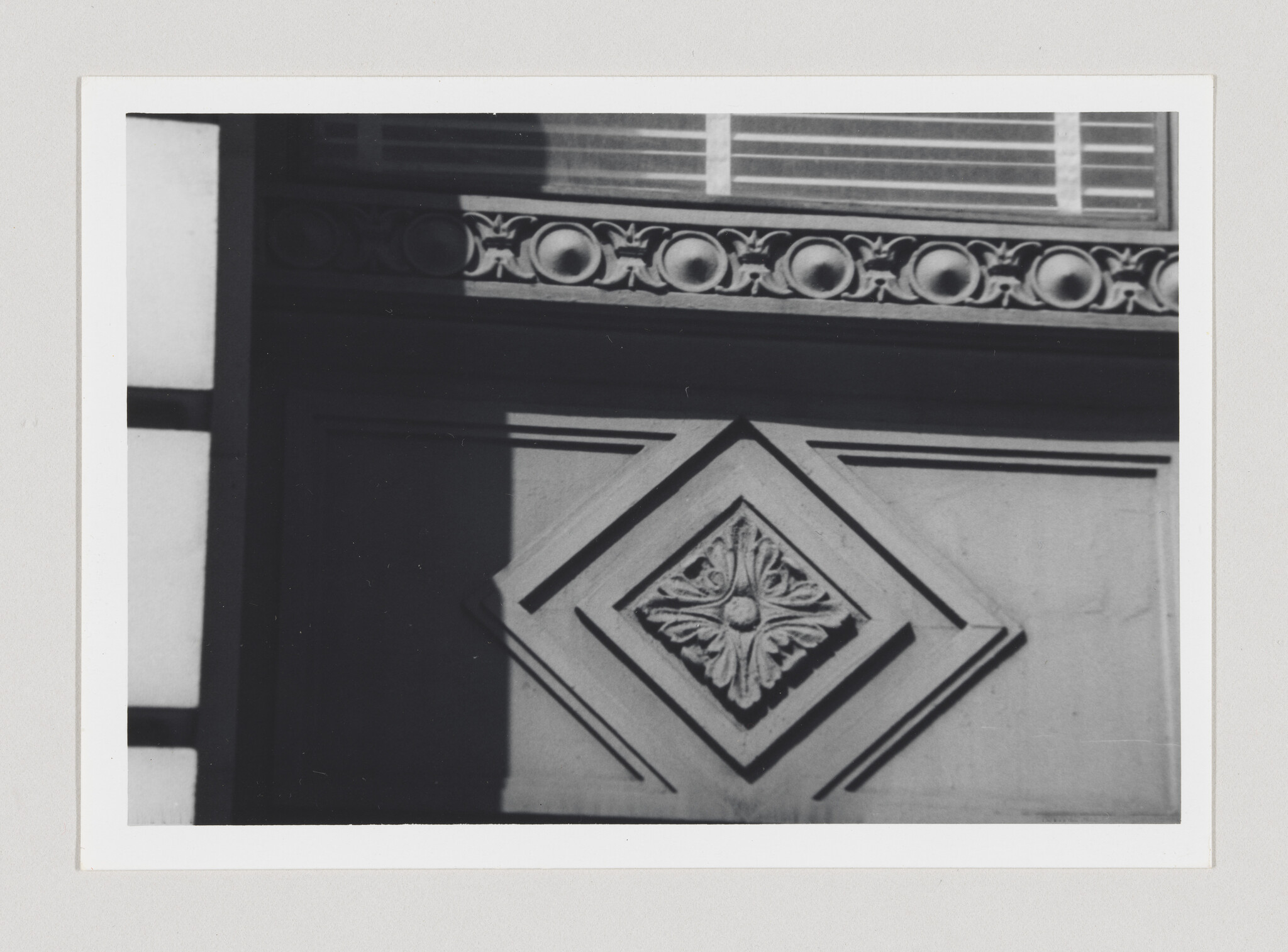 A black and white photo showing architectural details of a building, with a decorative frieze featuring circular patterns under a window and a diamond-shaped ornament with a floral design on the wall.