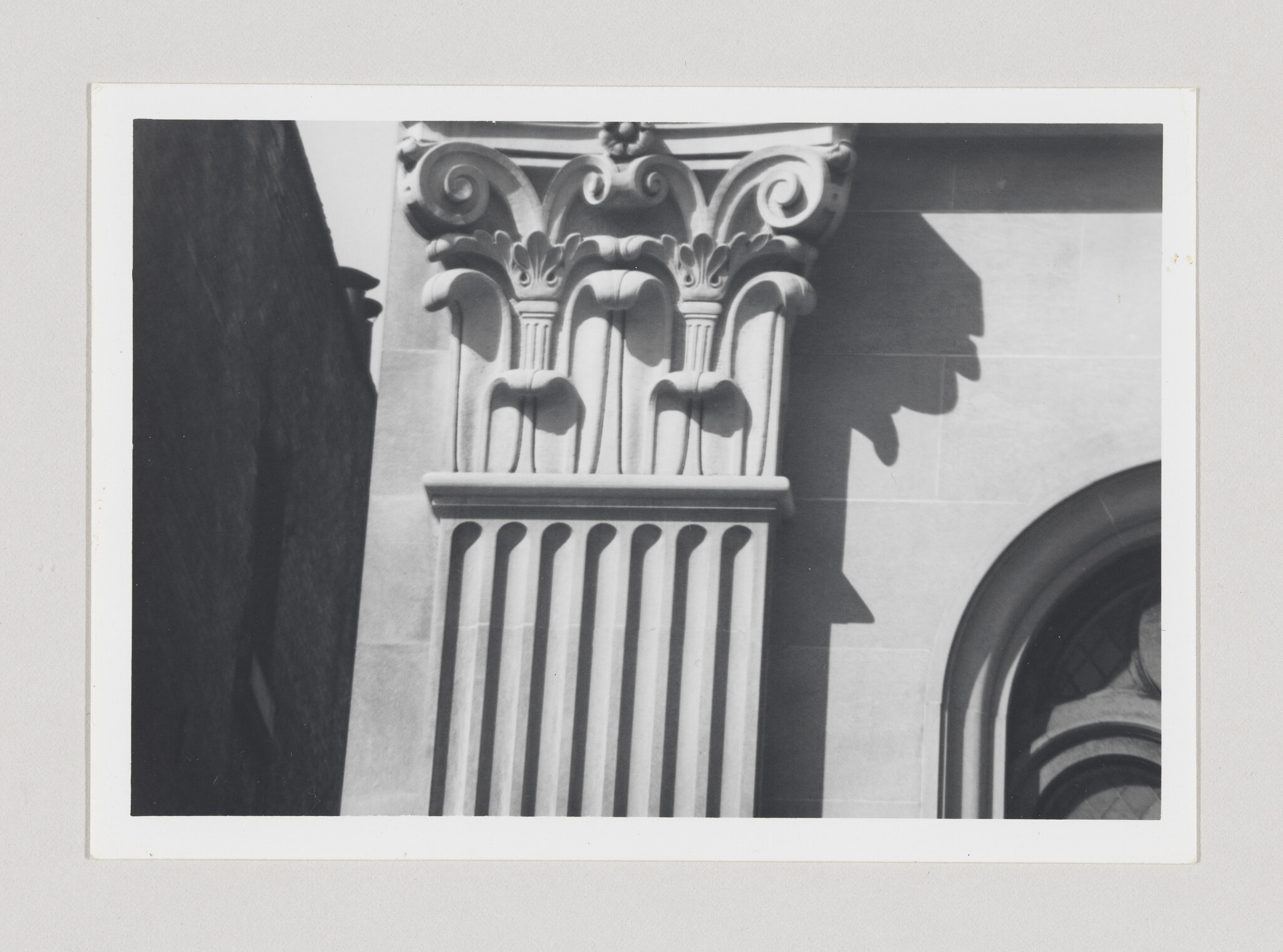 A black and white photograph of a classical architectural column with ornate Corinthian capital detailing, partially shadowed by an unseen structure, with a glimpse of an arched window to the right.