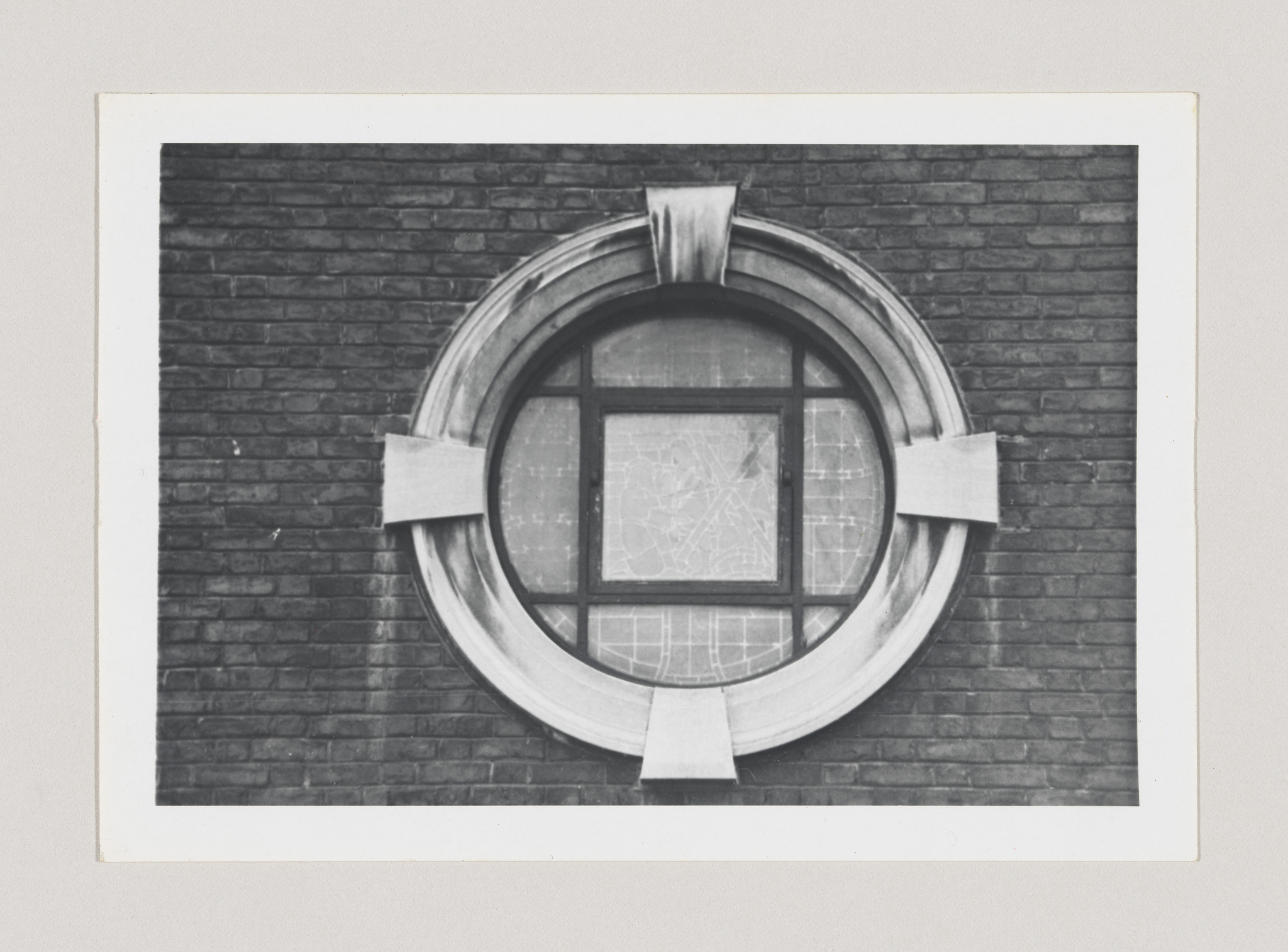 A black and white photograph of a round window with a unique geometric design set in a brick wall. The window features a prominent circular frame with a cross-shaped mullion pattern in the center.