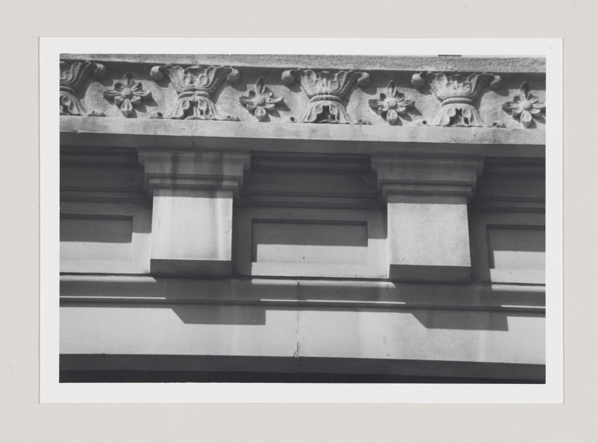 Stone building facade with decorative floral frieze and three projecting pilasters casting sharp shadows.