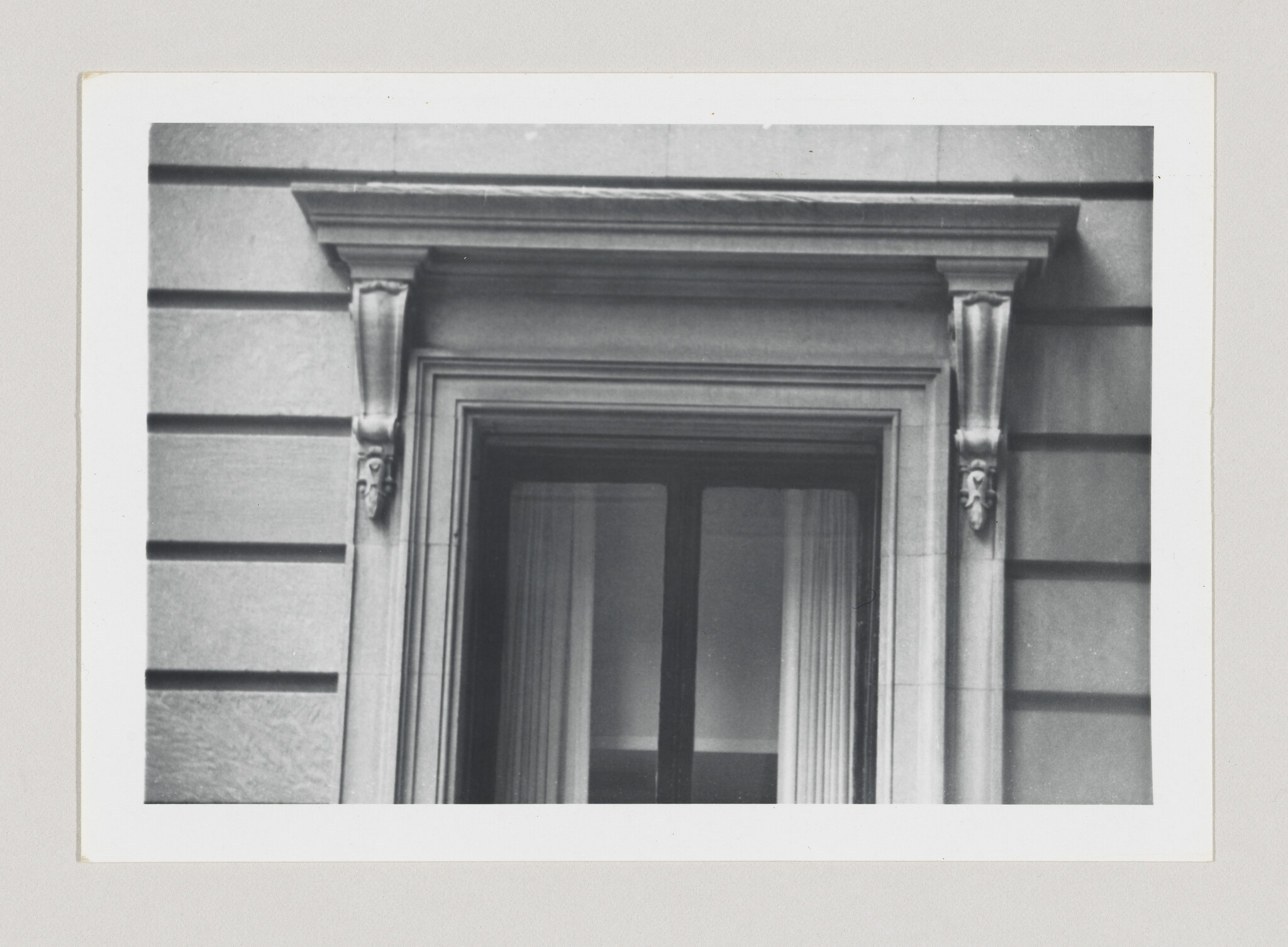 A classical stone window framed by decorative corbels and closed curtains behind glass.