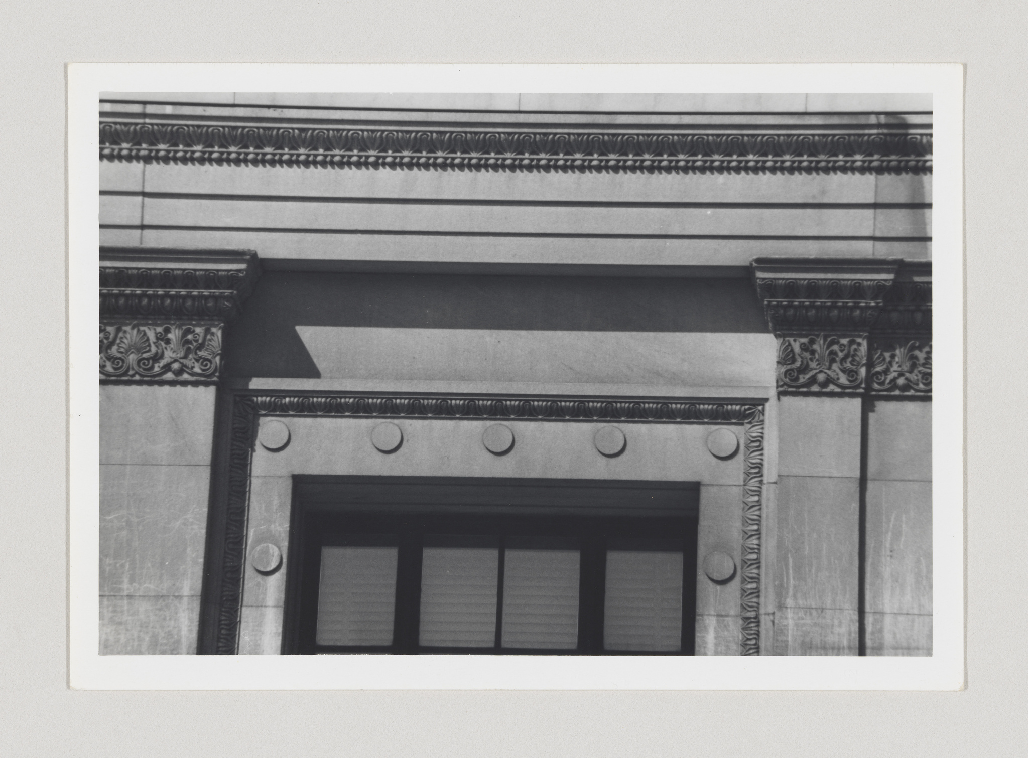 A black and white photograph of an architectural detail showing a section of a building facade with classical elements, including a cornice with dentil molding, a frieze with decorative relief, and a set of three windows with shutters framed by pilasters and topped with a lintel.