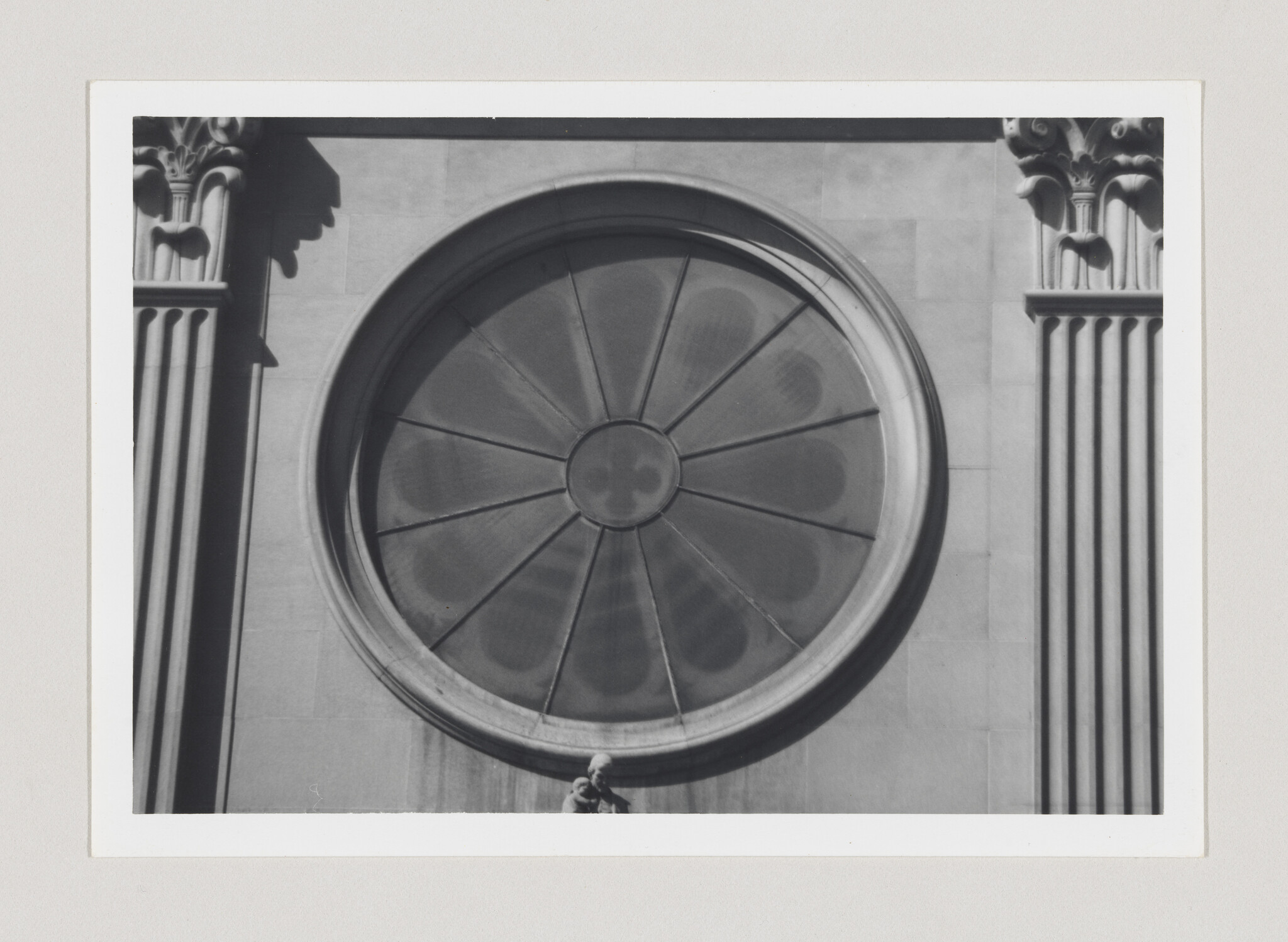 A large round stone window with radial panes sits between two fluted columns above a small statue.