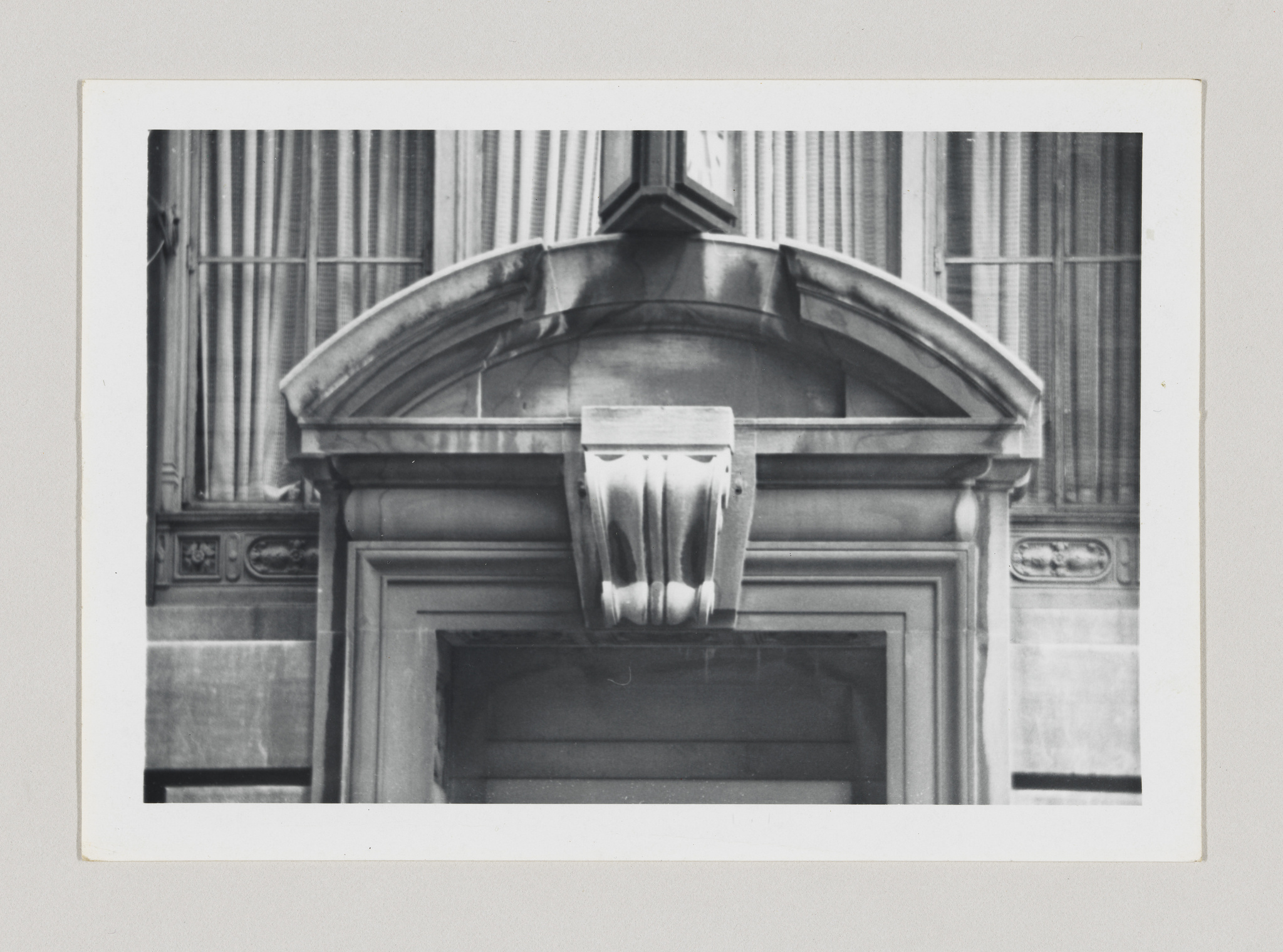 A black and white photograph of an architectural detail showing a classical stone window frame with a decorative cornice and a sculpted keystone above the window. The window is closed with blinds behind the glass, and the surrounding wall features ornate moldings. The photo is bordered by a white edge, suggesting it is a print.