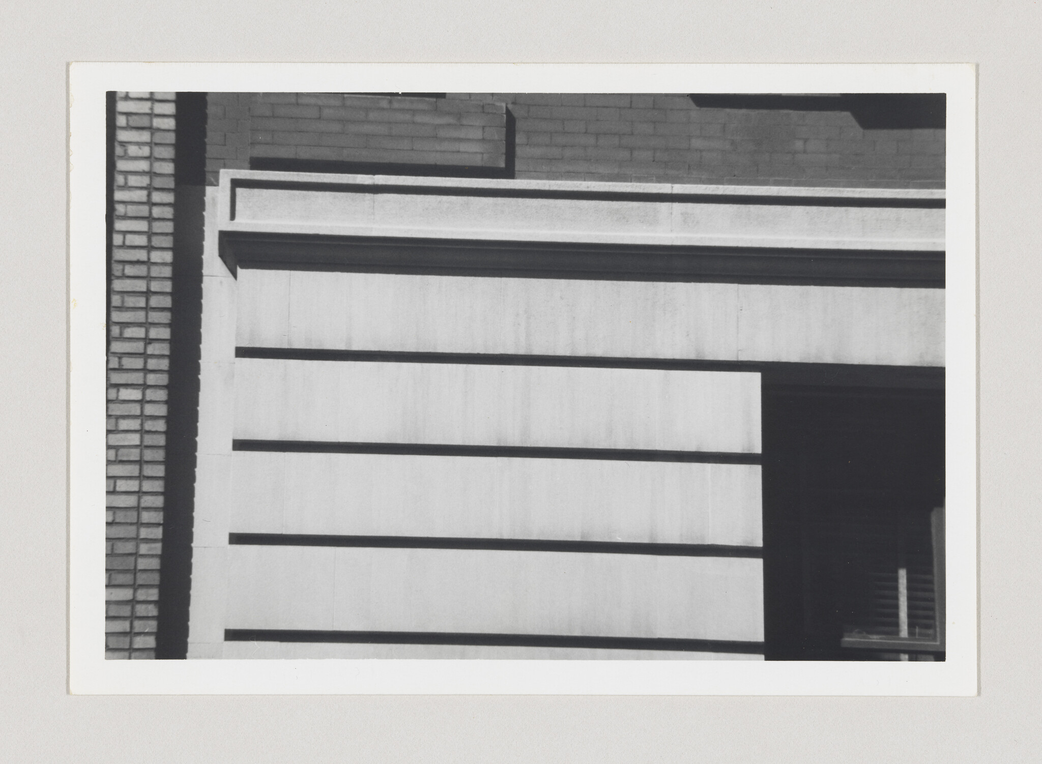 Building facade showing horizontal concrete panels, a brick column on the left, and a dark window opening.