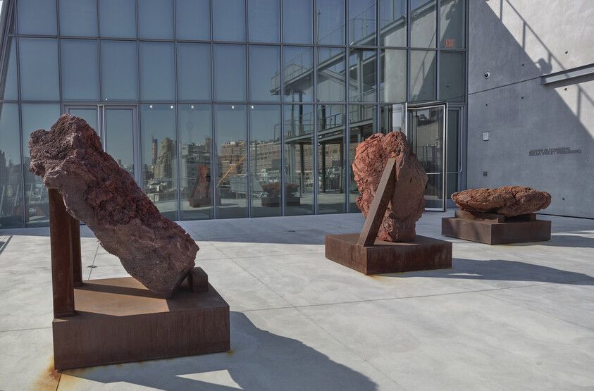 Three large rust-colored rock sculptures rest on metal bases outside a modern glass building.