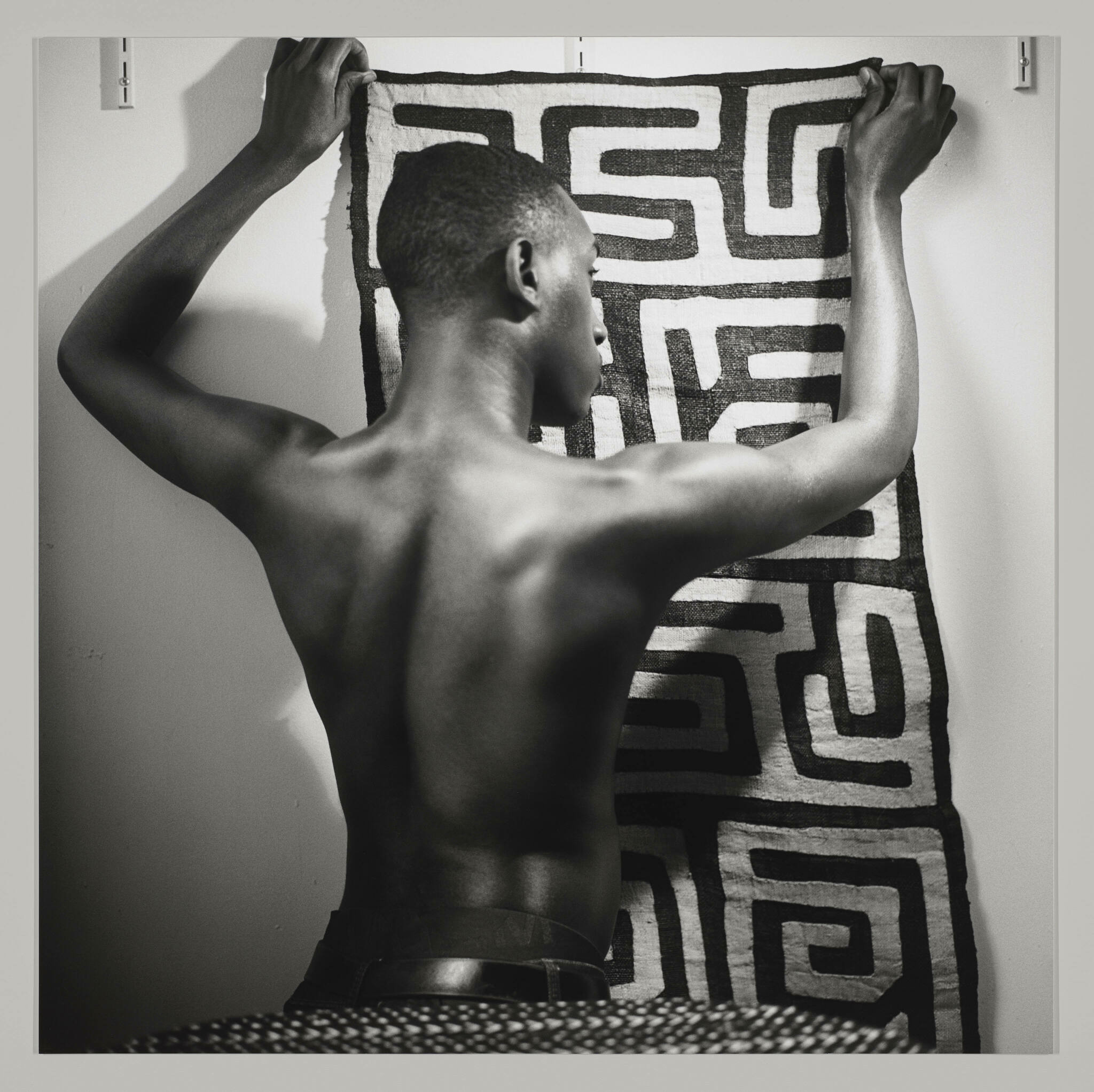 With their back to the camera, a shirtlless Black person with short hair holds a pattered cloth against a wall.