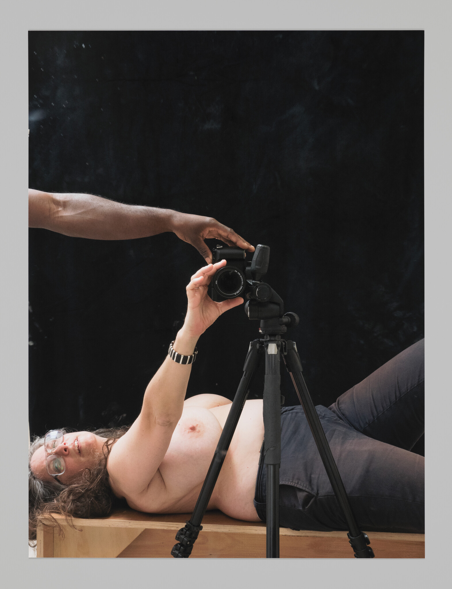 A topless person lies on a wooden box taking a self-portrait with a camera on a tripod.