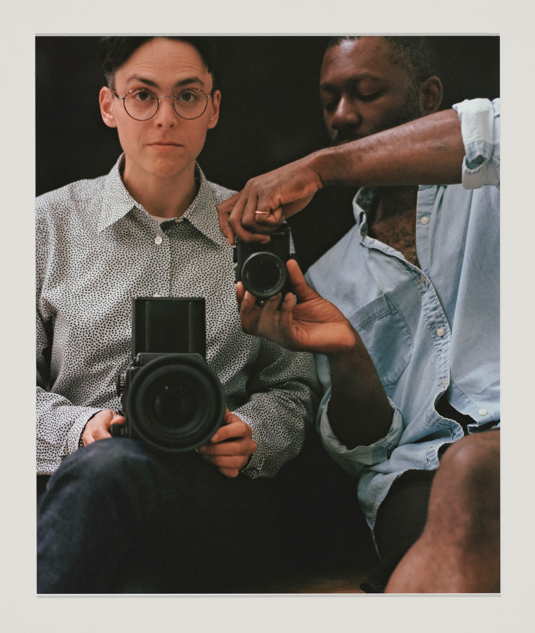 Two seated people, a white person in glasses and a Black person with a mustache, photograph their reflections with different size cameras
