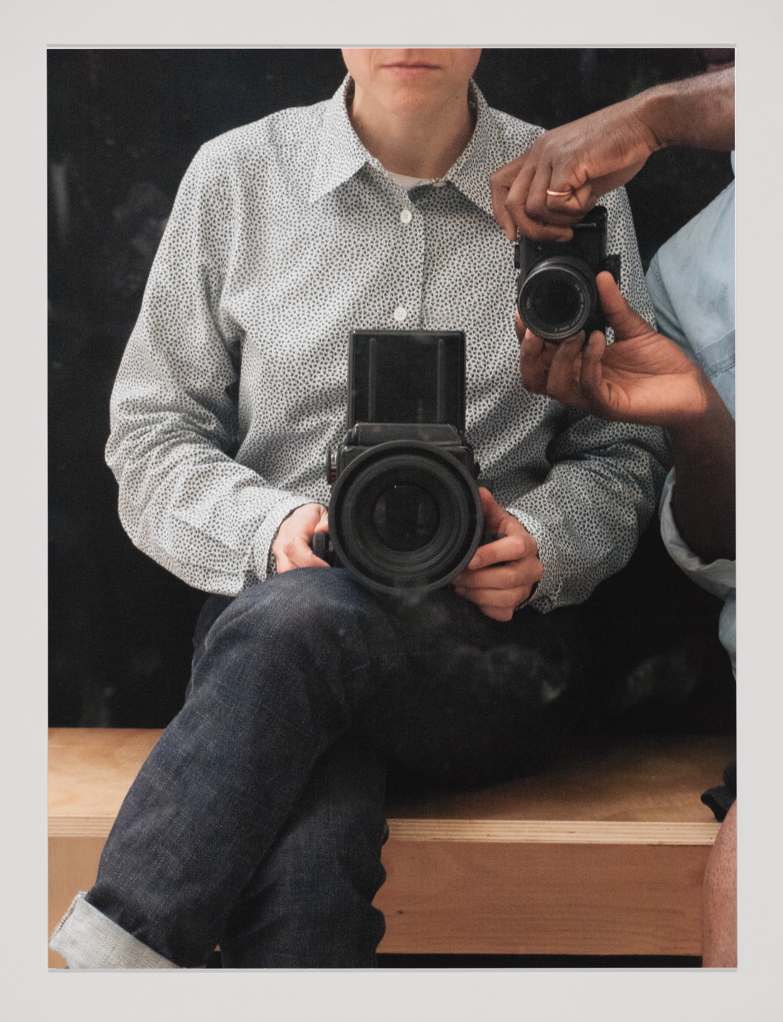 A seated white person holds a large camera in their lap, their face cropped, and a Black person reaches into the frame holding a smaller camera
