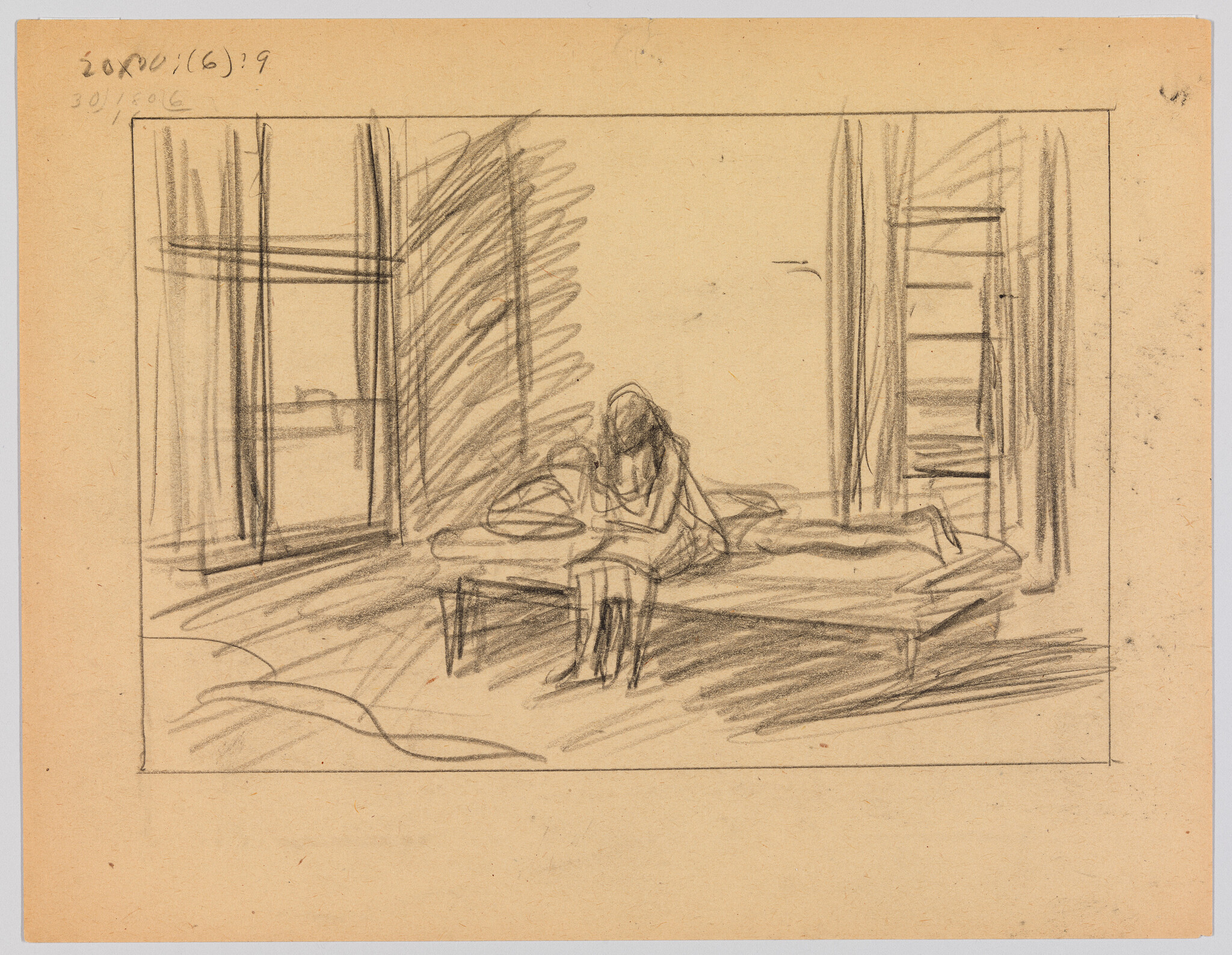 A person sits on the edge of a bed by tall windows, leaning forward and holding something.