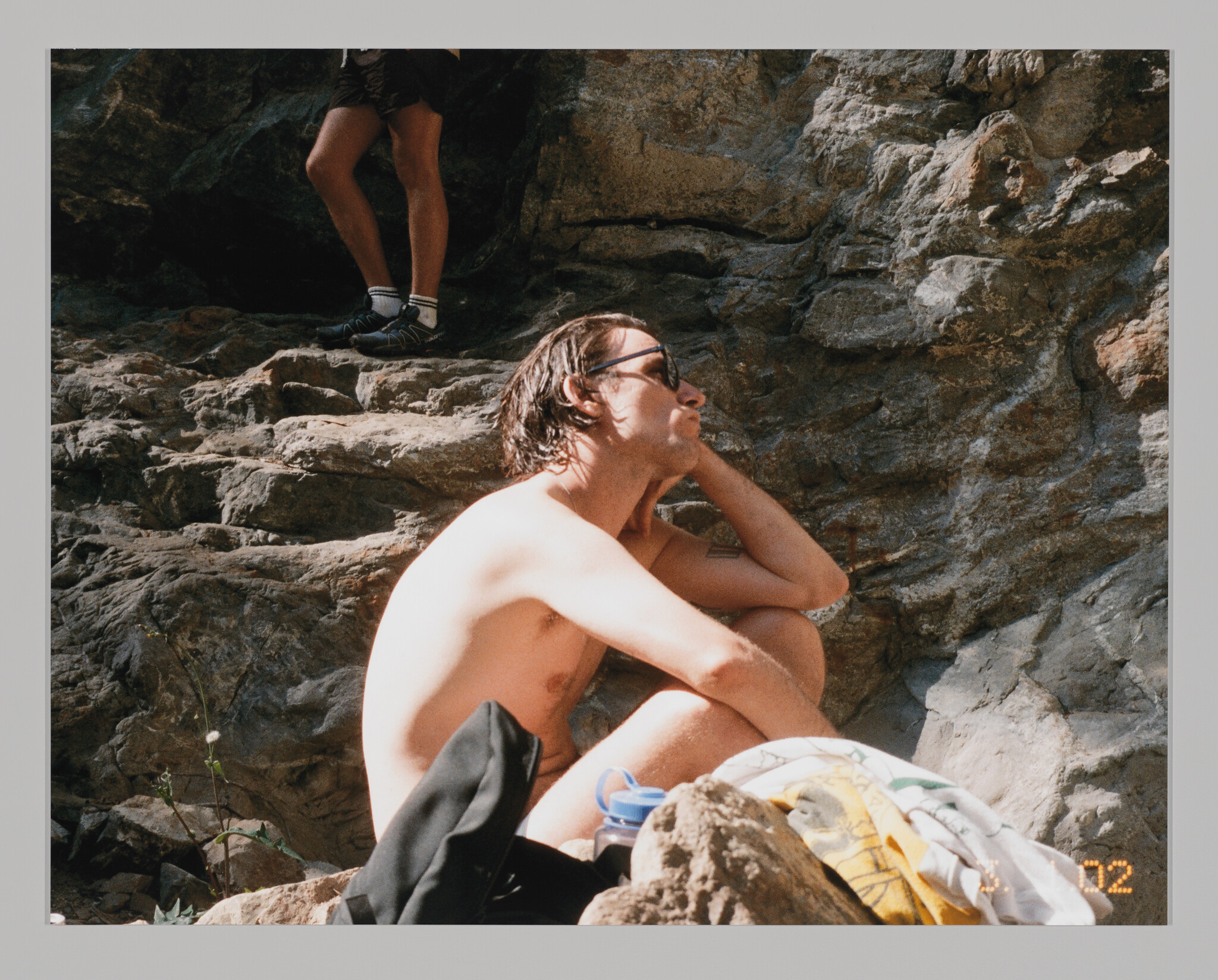 A shirtless man wearing sunglasses sits on rocks, resting his chin on his hand while relaxing.