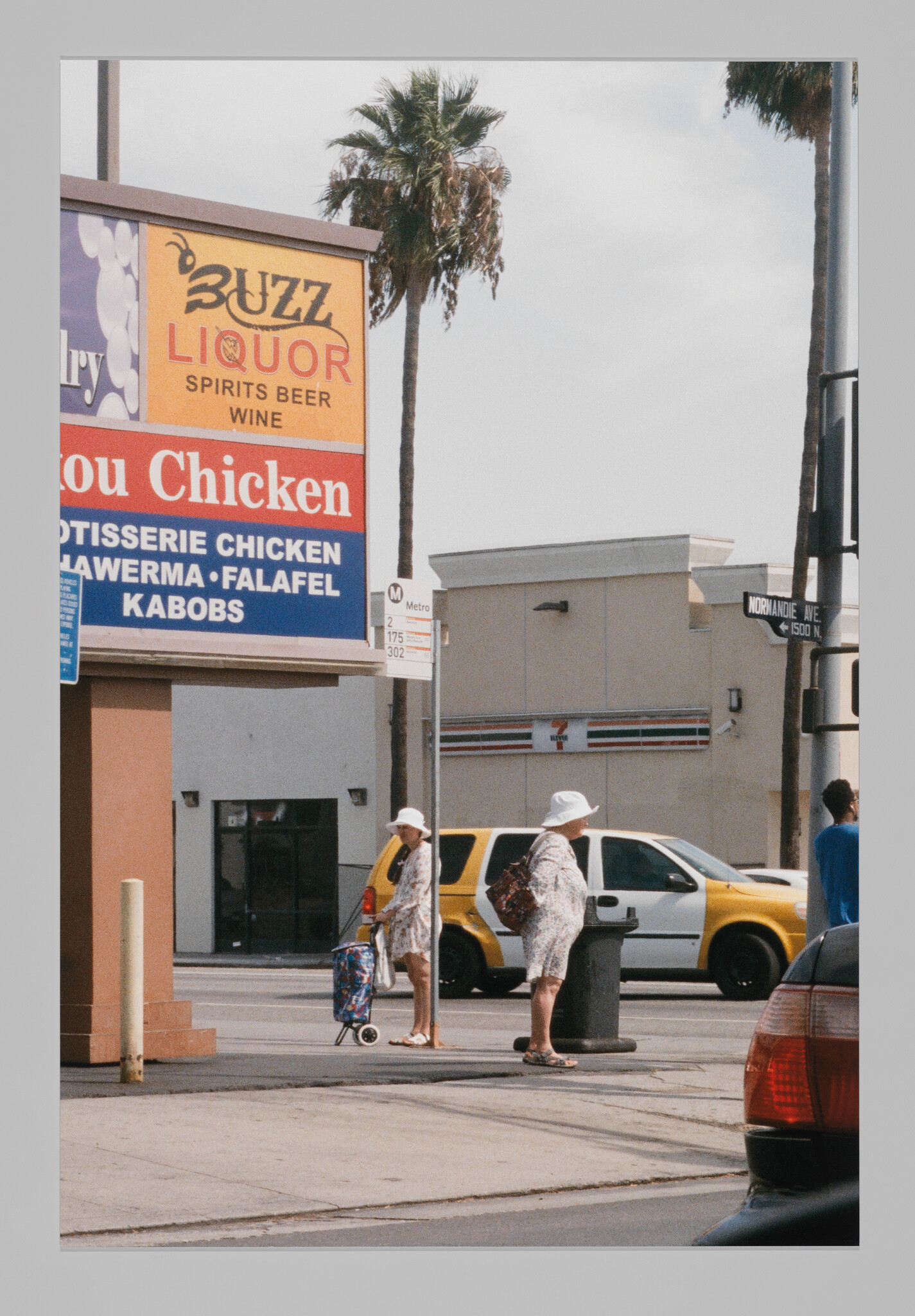 Two older women in white hats wait at a sunny street corner near a liquor store sign.