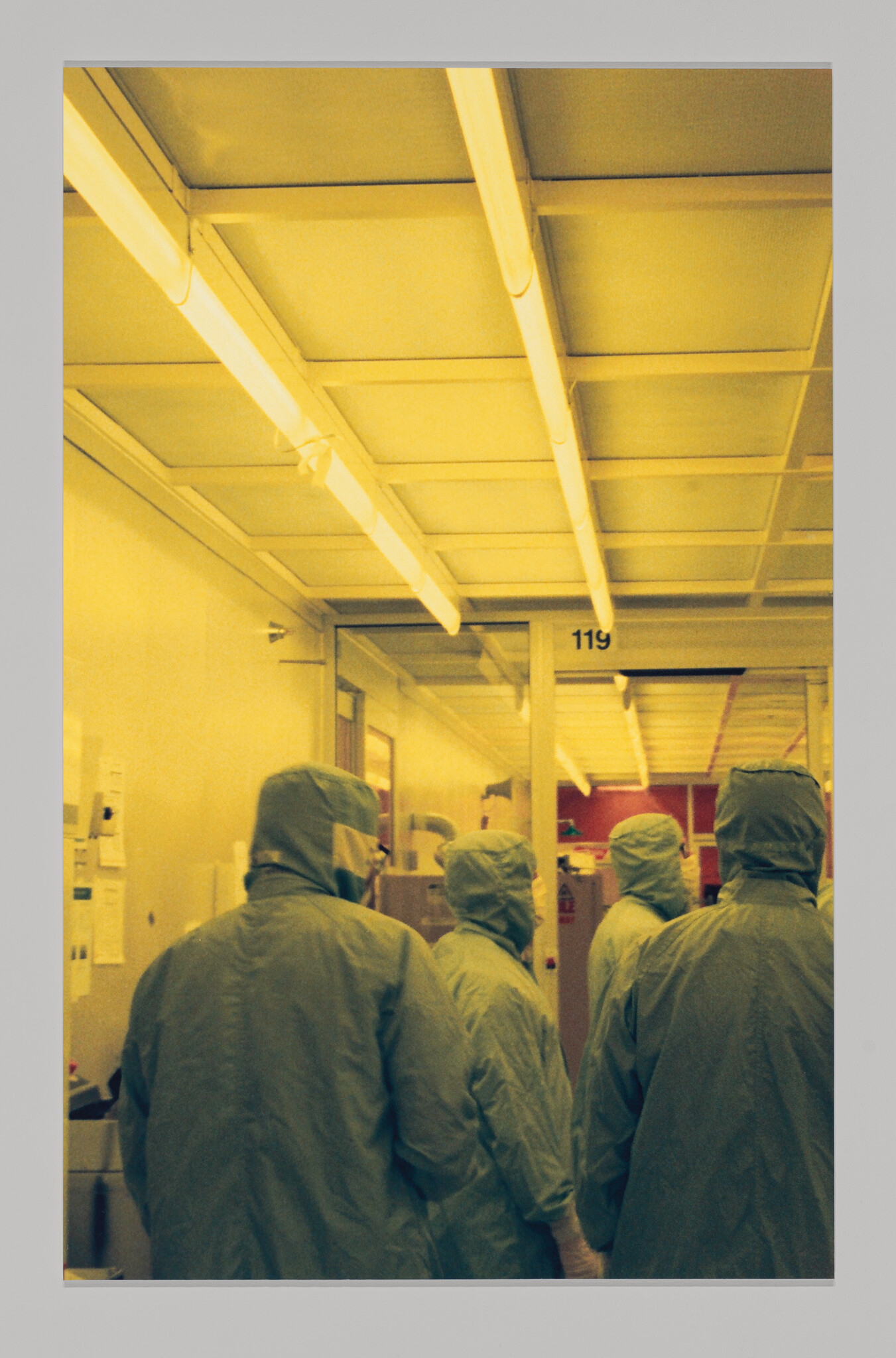 Four people in protective suits stand together in a yellow-lit cleanroom corridor.