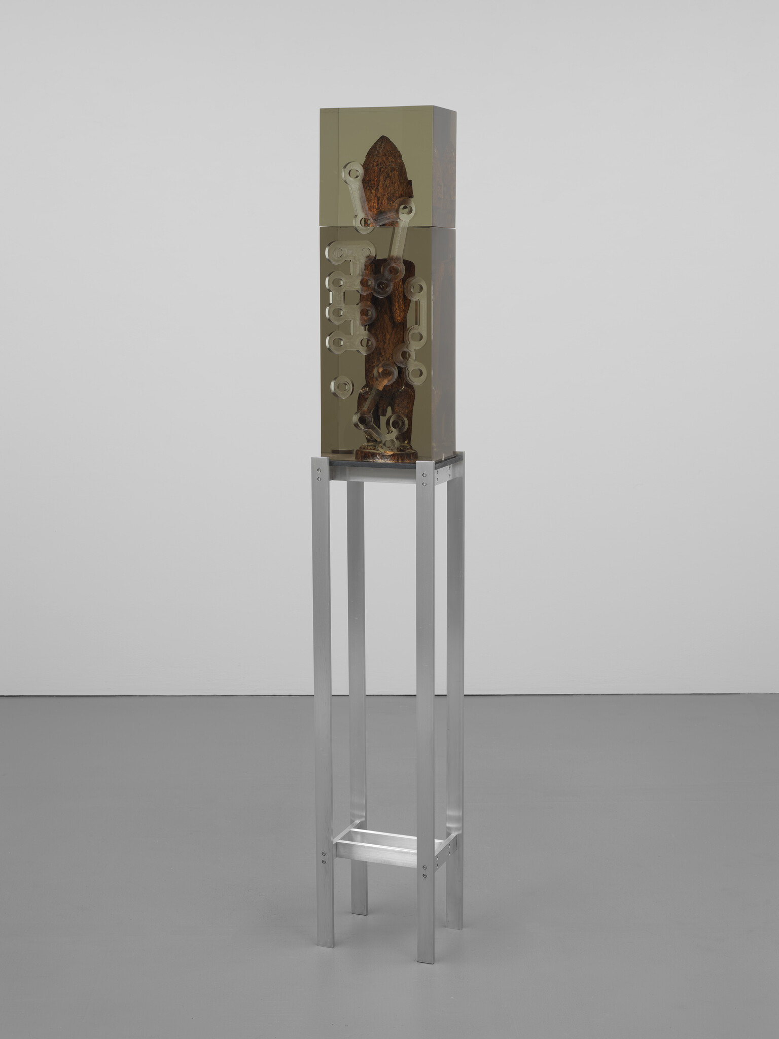 A tall metal stand holds a clear rectangular case with a wooden object and visible gears.