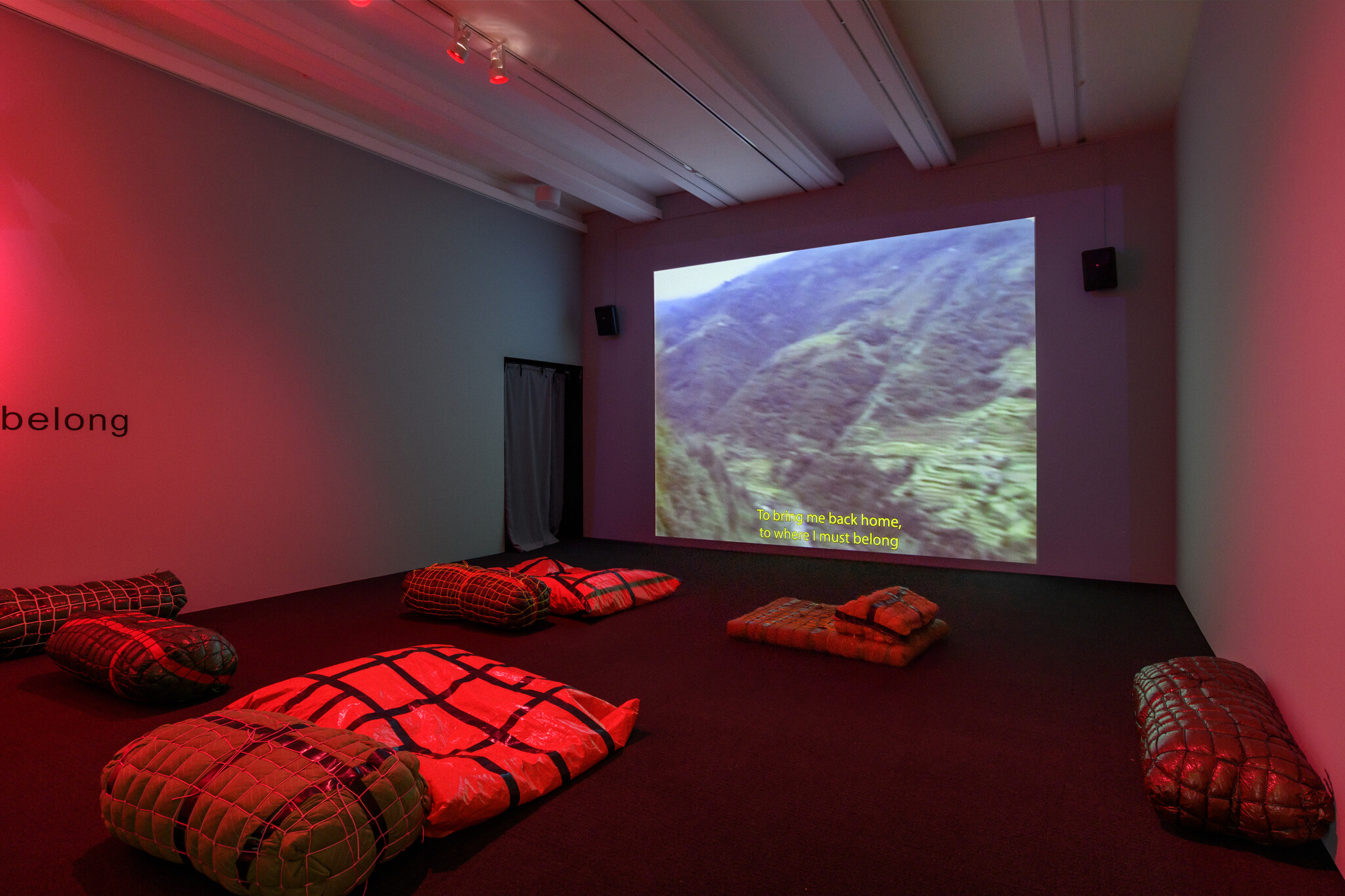 Room with red lighting, cushions on the floor, and a video projection of a landscape with subtitles.